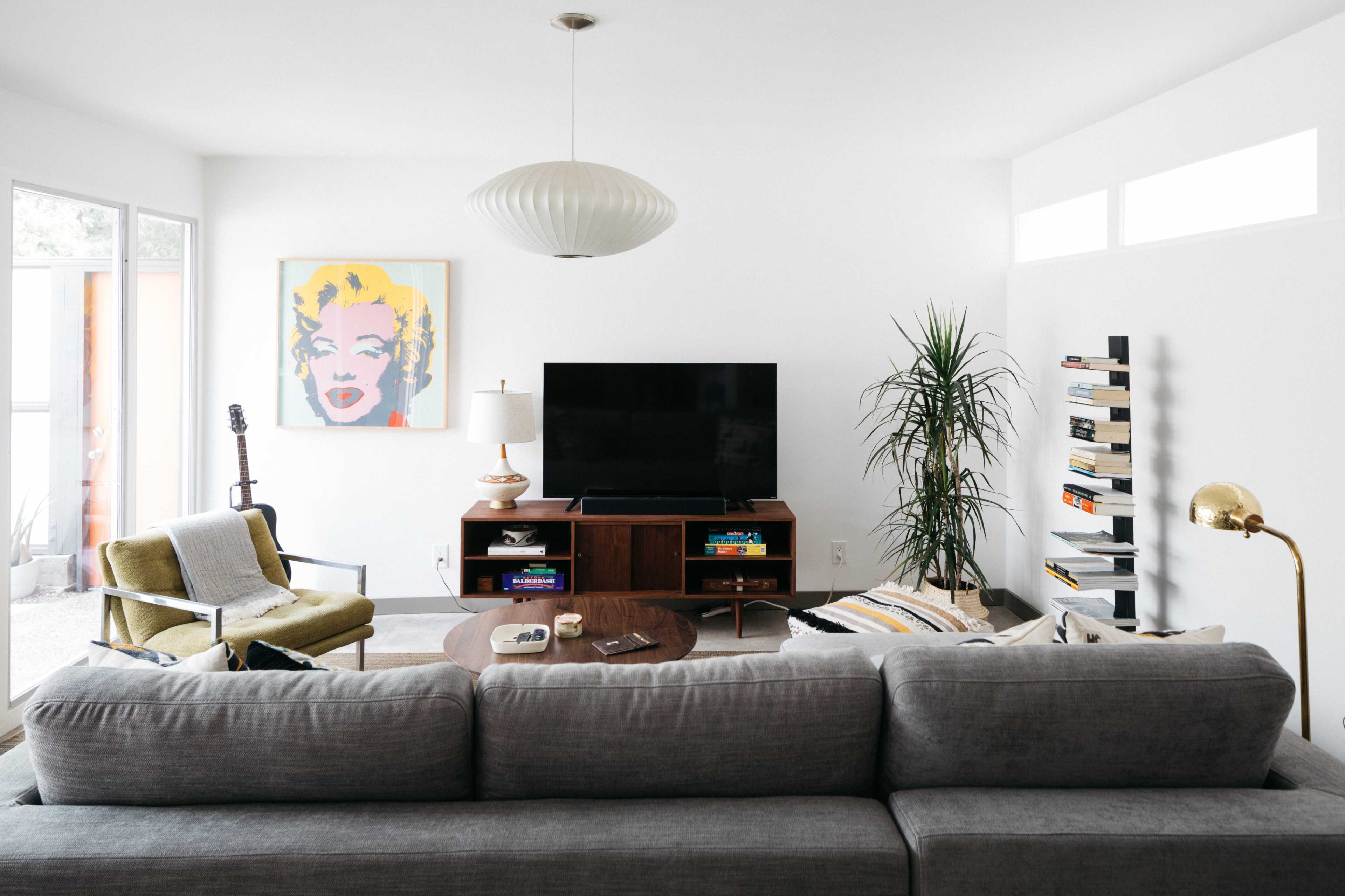 A modern living room features a gray sectional sofa, a brown wooden TV stand with a flat-screen TV, a guitar resting nearby, and a painting of a woman on the wall.