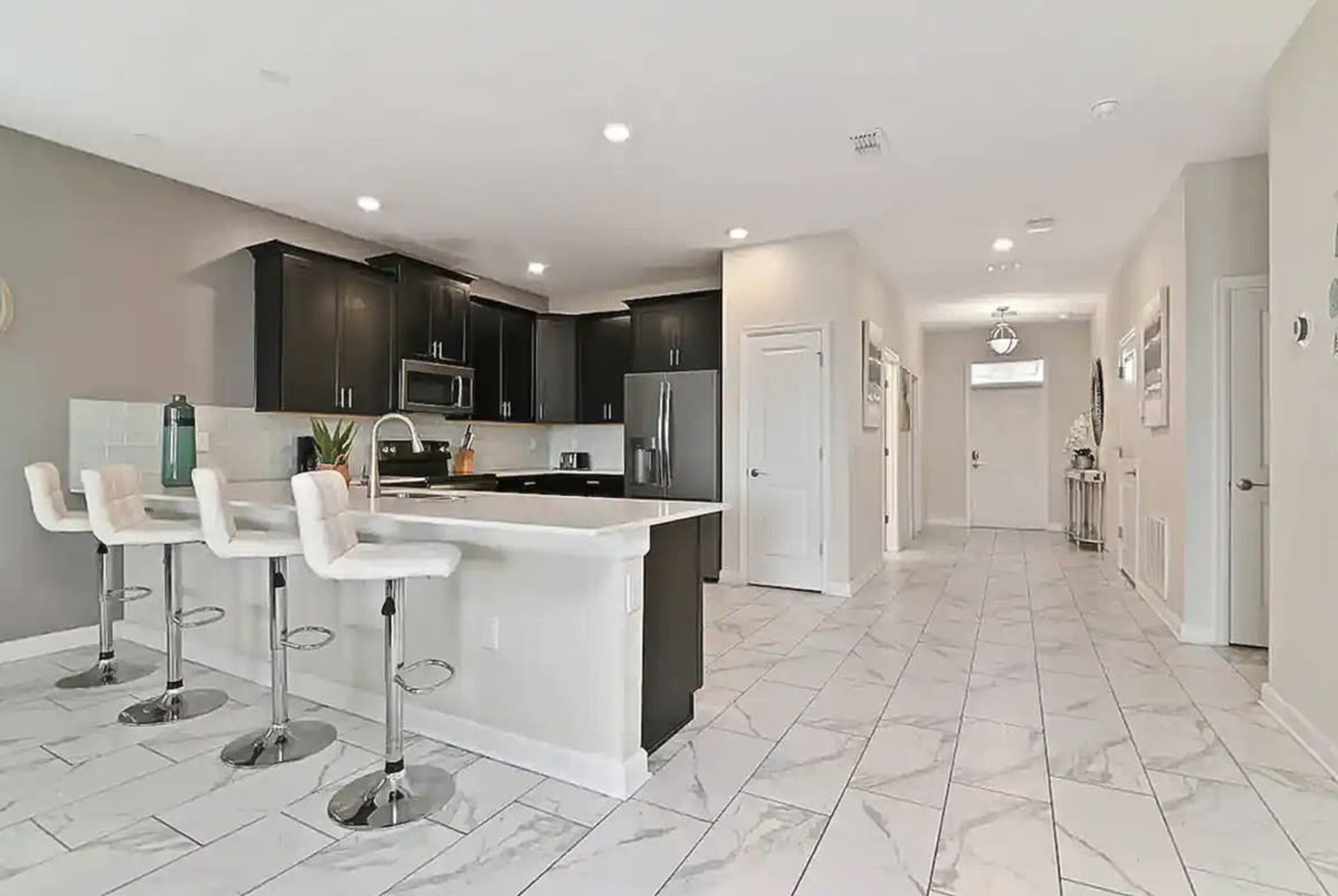 A modern kitchen area with black cabinets, a white island, and bar stools, leading into a hallway with light-colored walls and tile flooring.