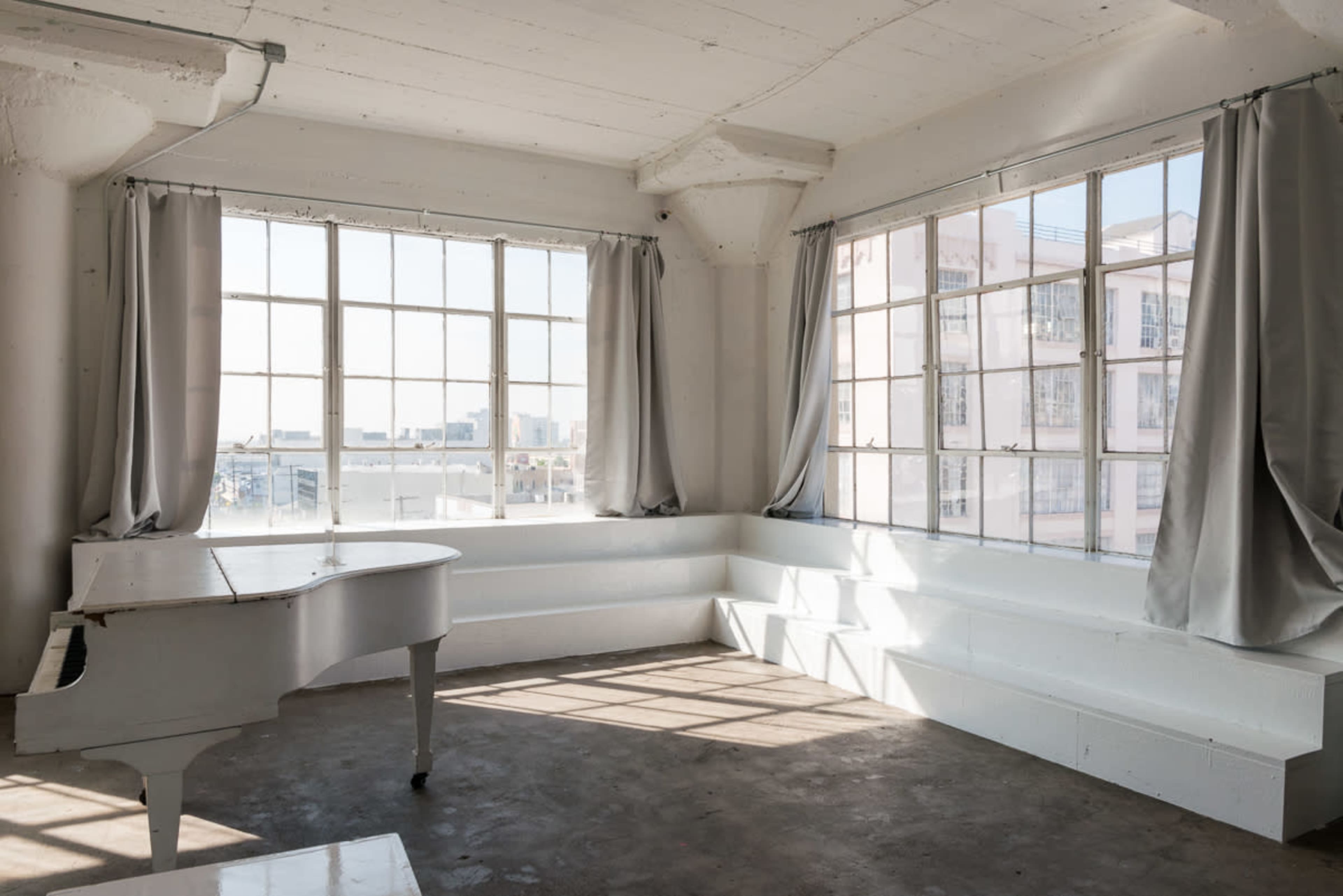 A bright, minimalist room features large windows with sheer curtains, a white grand piano, and built-in seating along the walls.