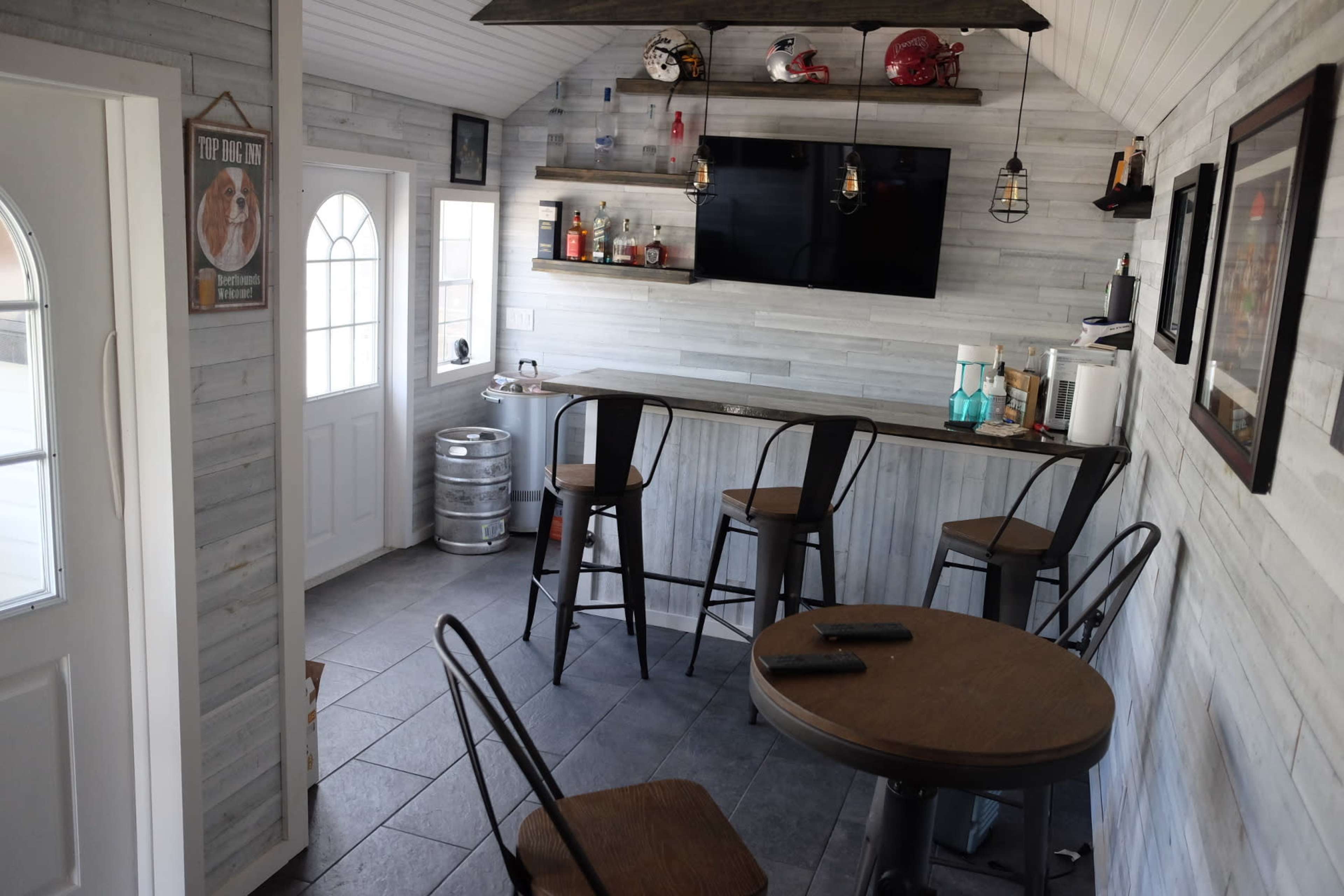 The interior features a small bar area with a television, several stools, and a round table, all set against a wall lined with bottles and decorative items.