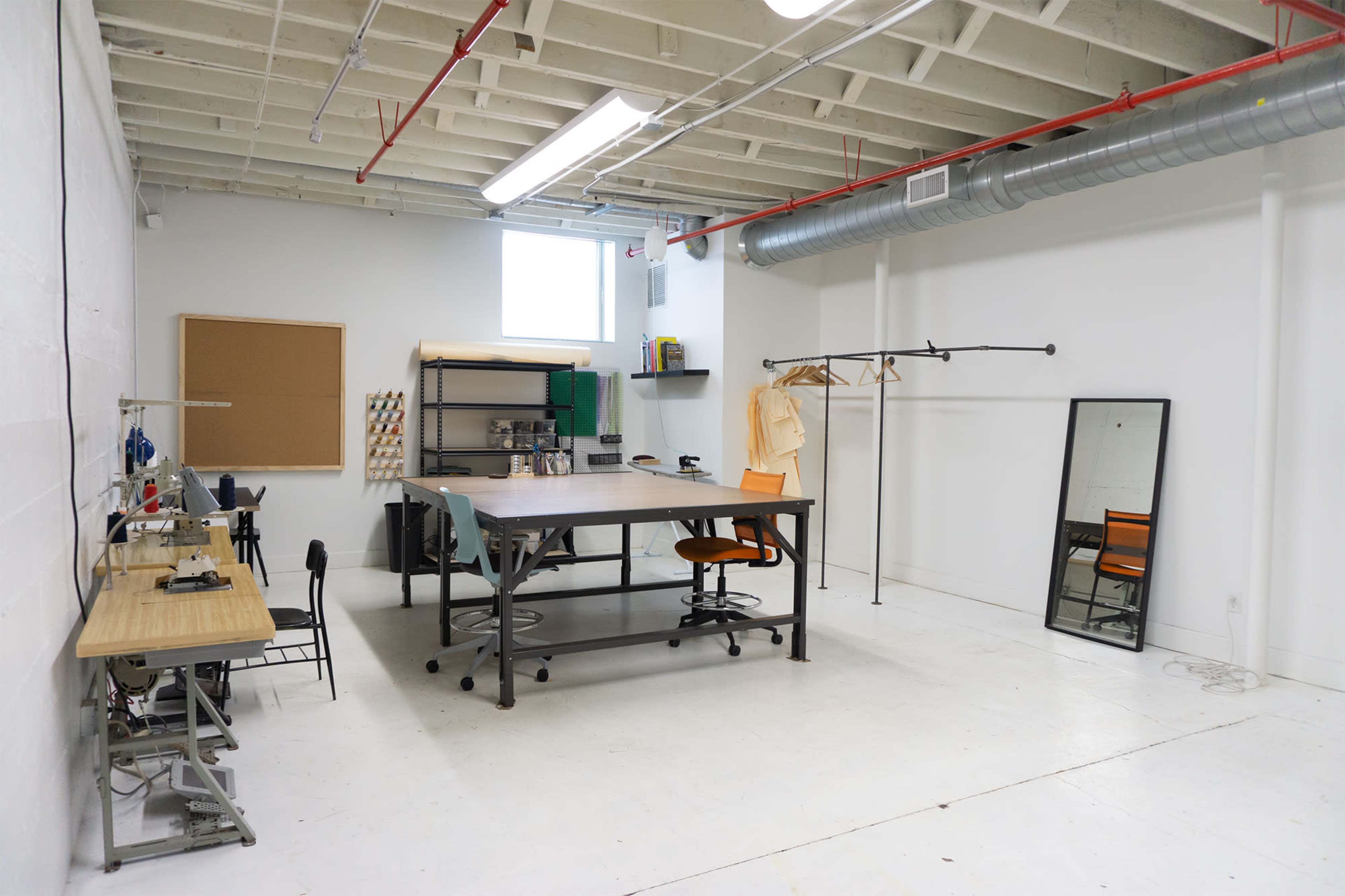 A spacious, industrial-style office workspace with a large central table, chairs, a clothes rack, and a mirror against a white wall.