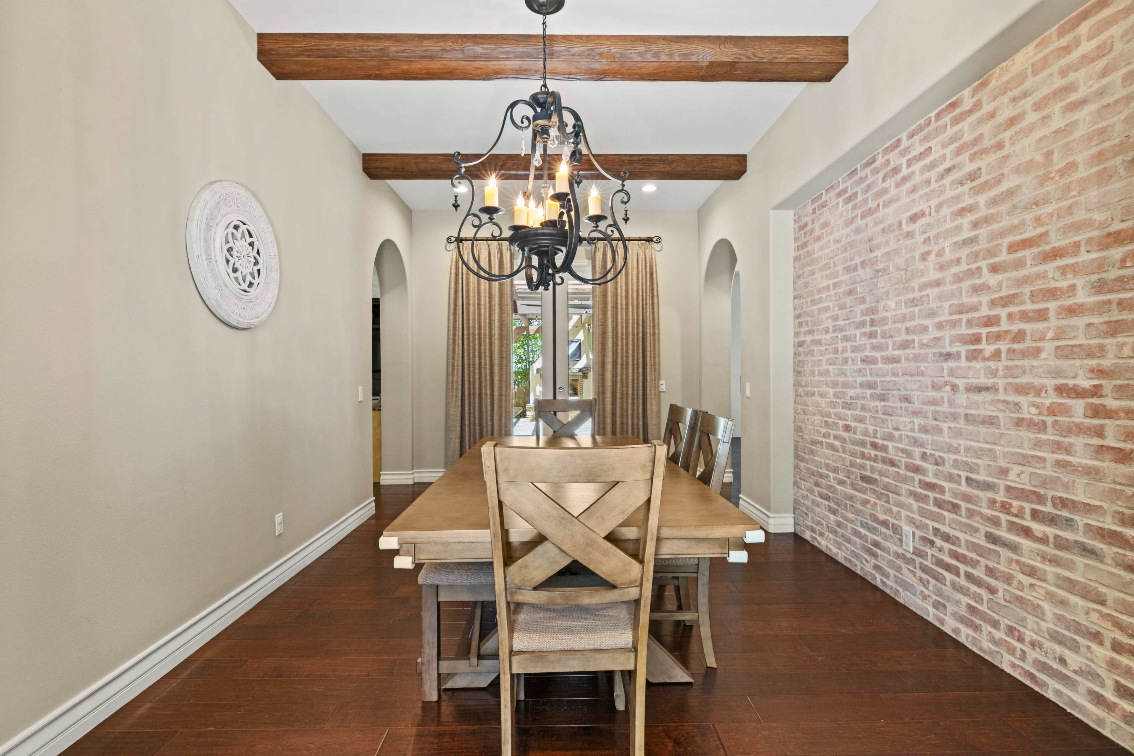 A dining room features a long wooden table with chairs, illuminated by a chandelier, against a brick wall and arched openings.