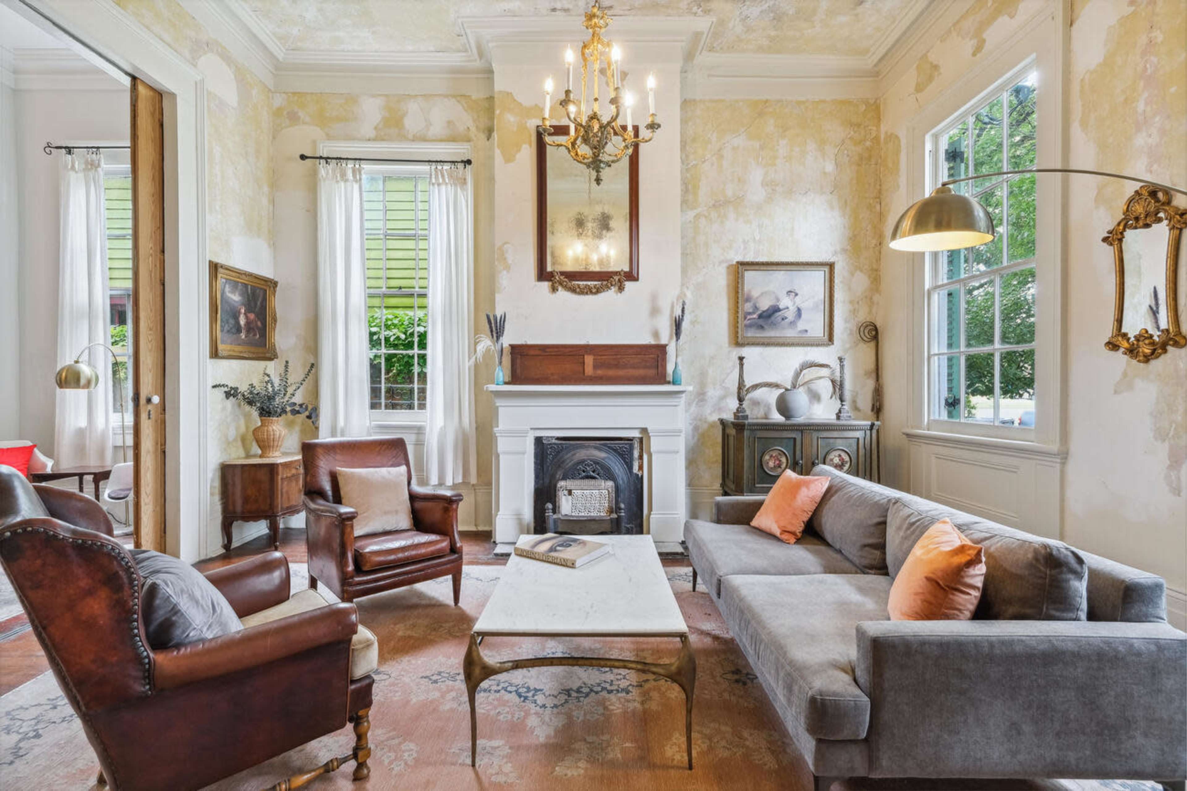 A living room with a chandelier, a fireplace, two armchairs, and a sofa, surrounded by faded wallpaper and large windows.