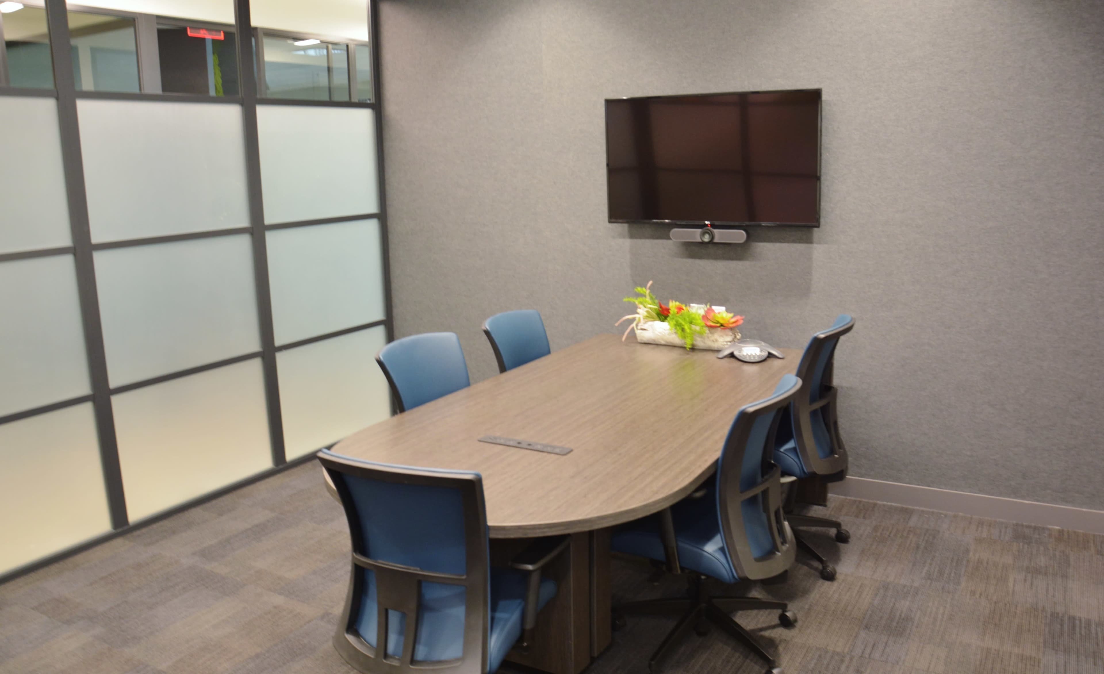 A conference room features a wooden table surrounded by six blue chairs, with a television mounted on the wall and a flower arrangement on the table.
