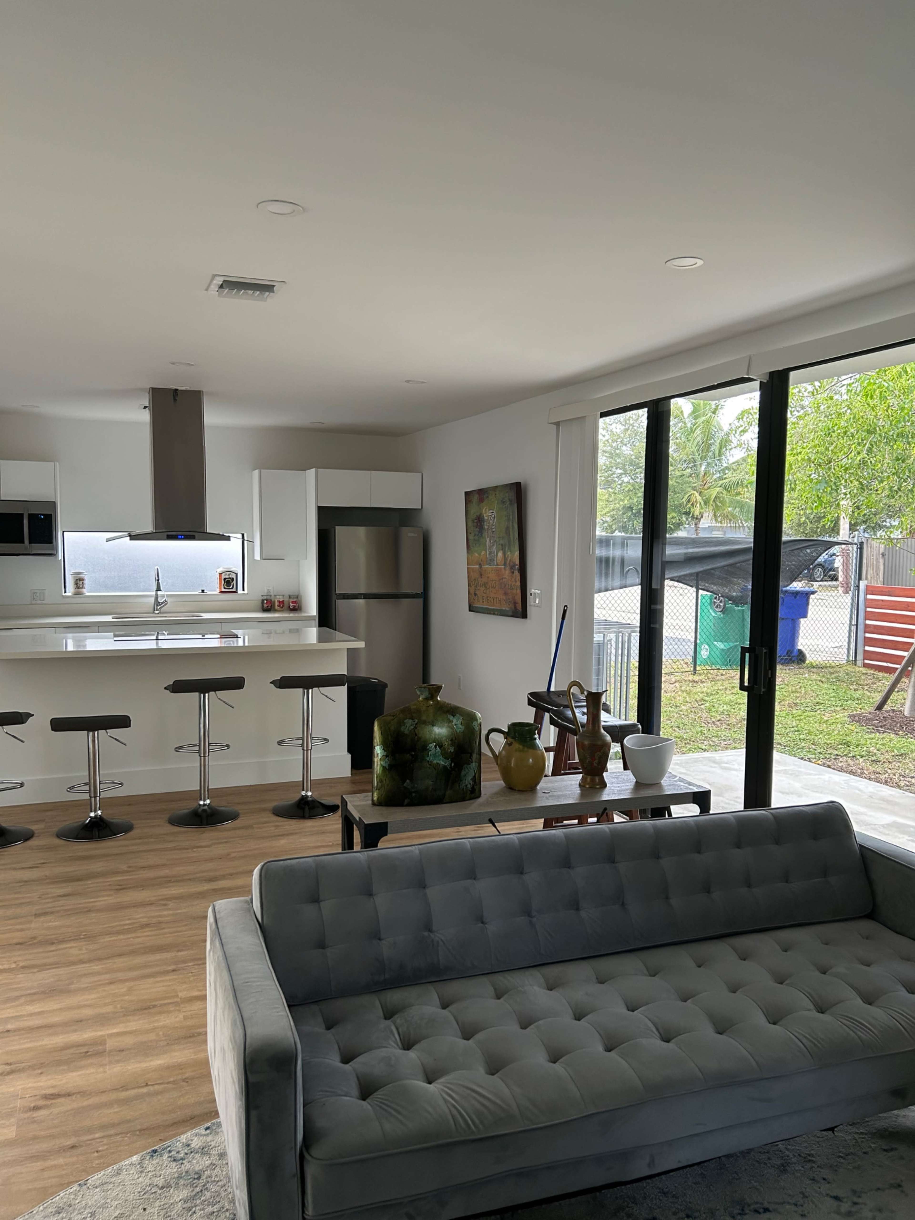 The image shows a modern kitchen and living area with a grey sofa, a kitchen island with bar stools, and large windows overlooking a yard.