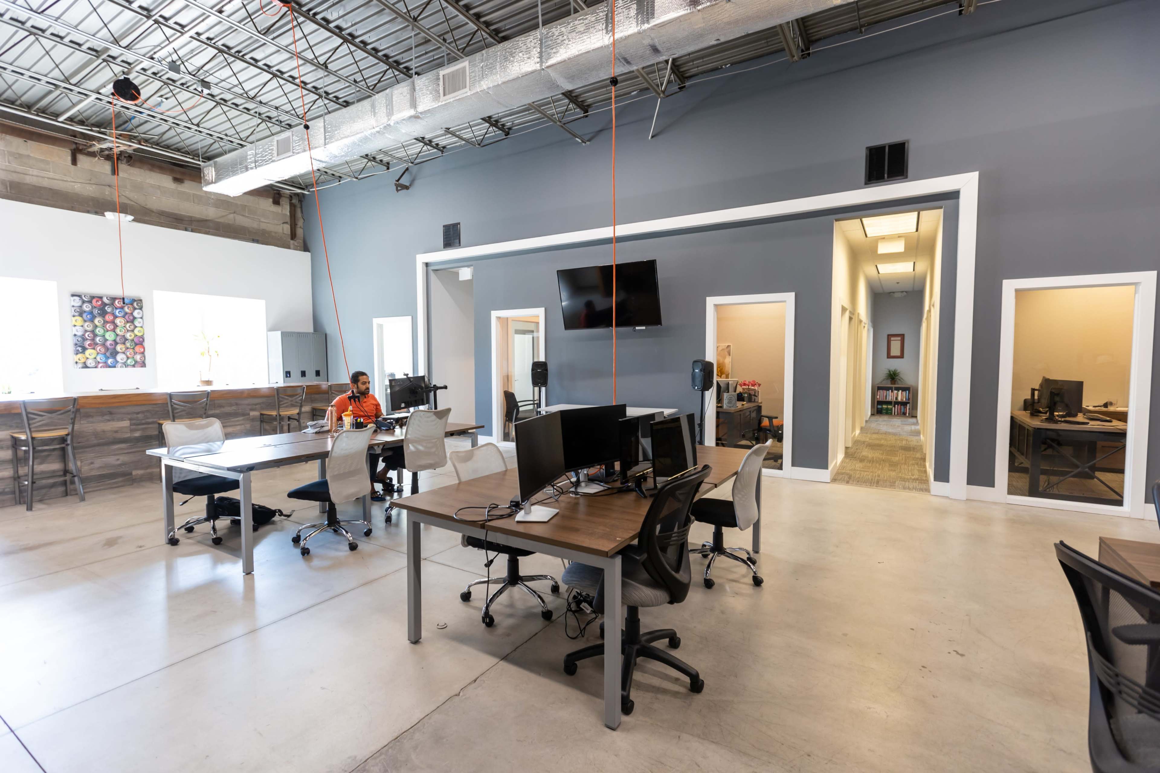 The image shows a modern, open-plan office space with several desks, computers, and a couple of employees working.