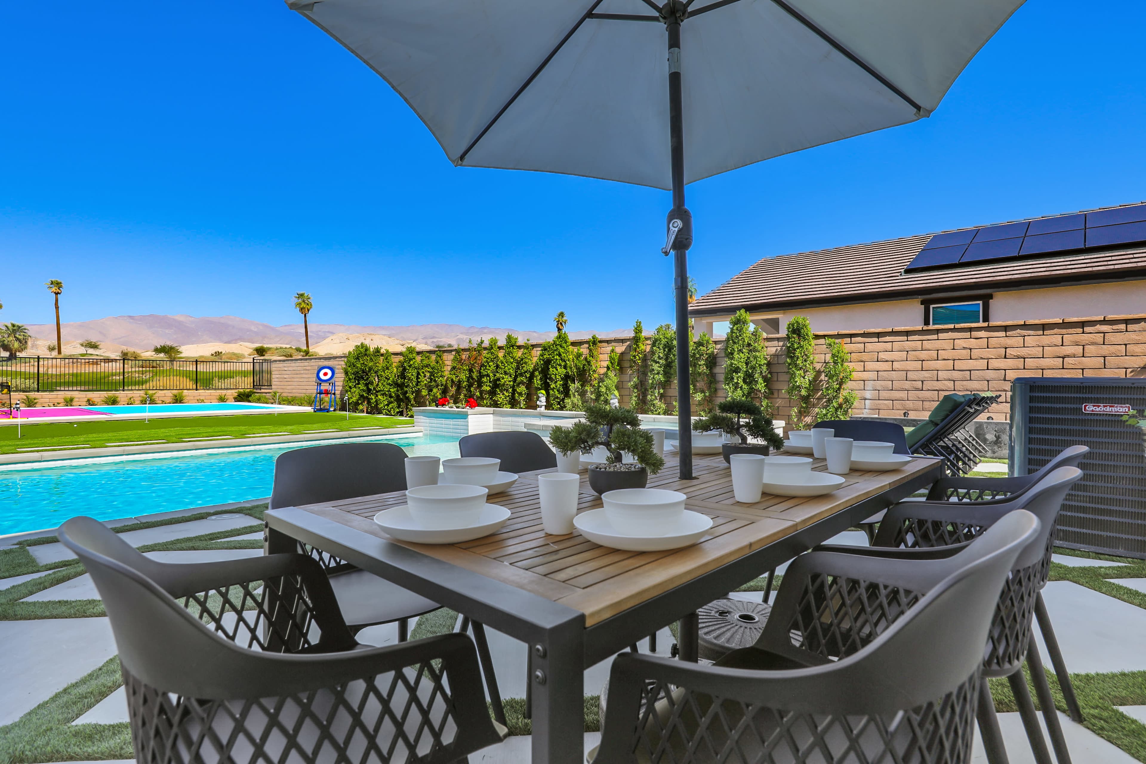A wooden table set with white dishes and cups is shaded by a large umbrella next to a swimming pool and landscaped yard.