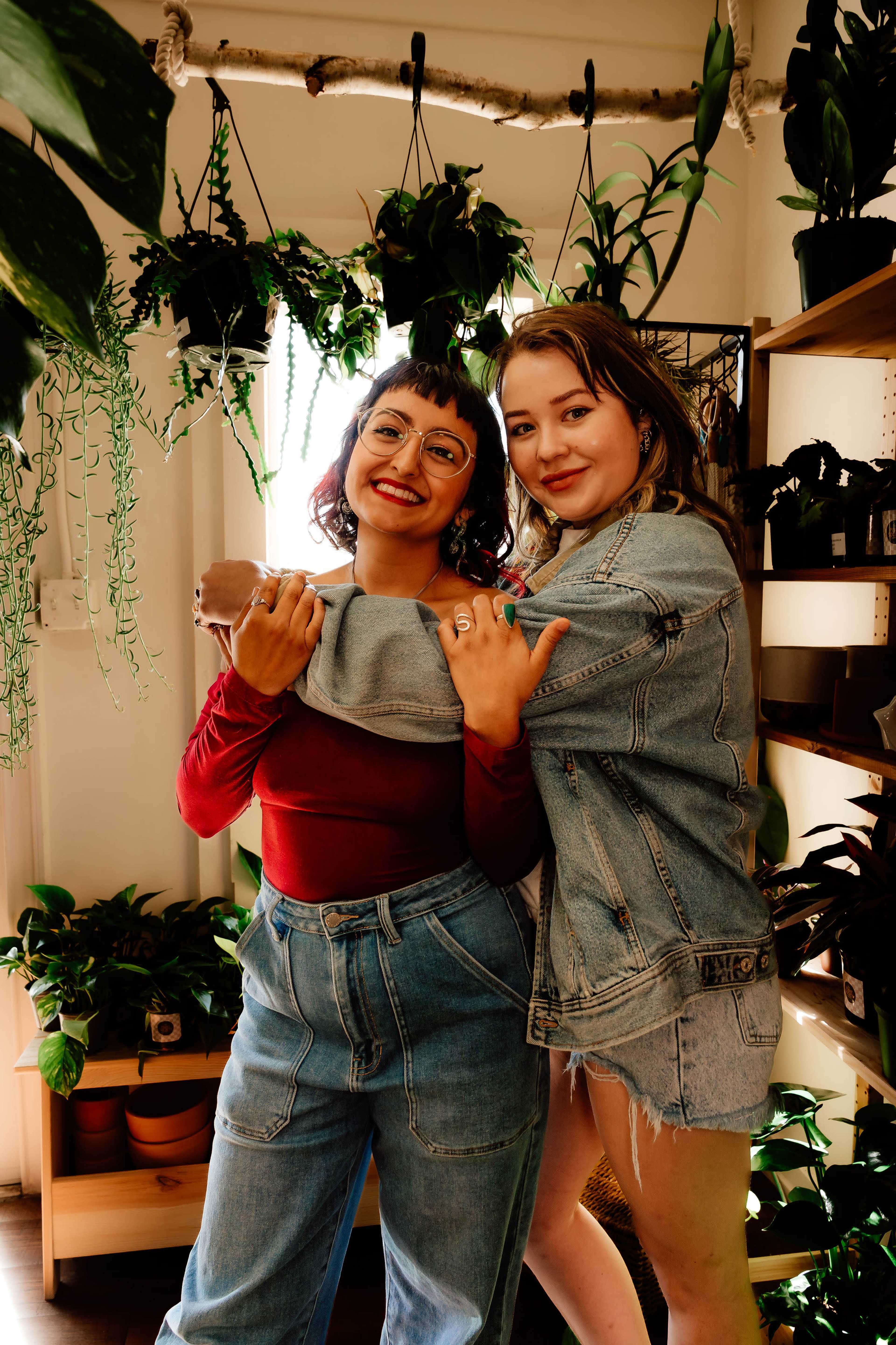 Two individuals pose together in a room filled with various potted plants, smiling and wearing casual outfits.