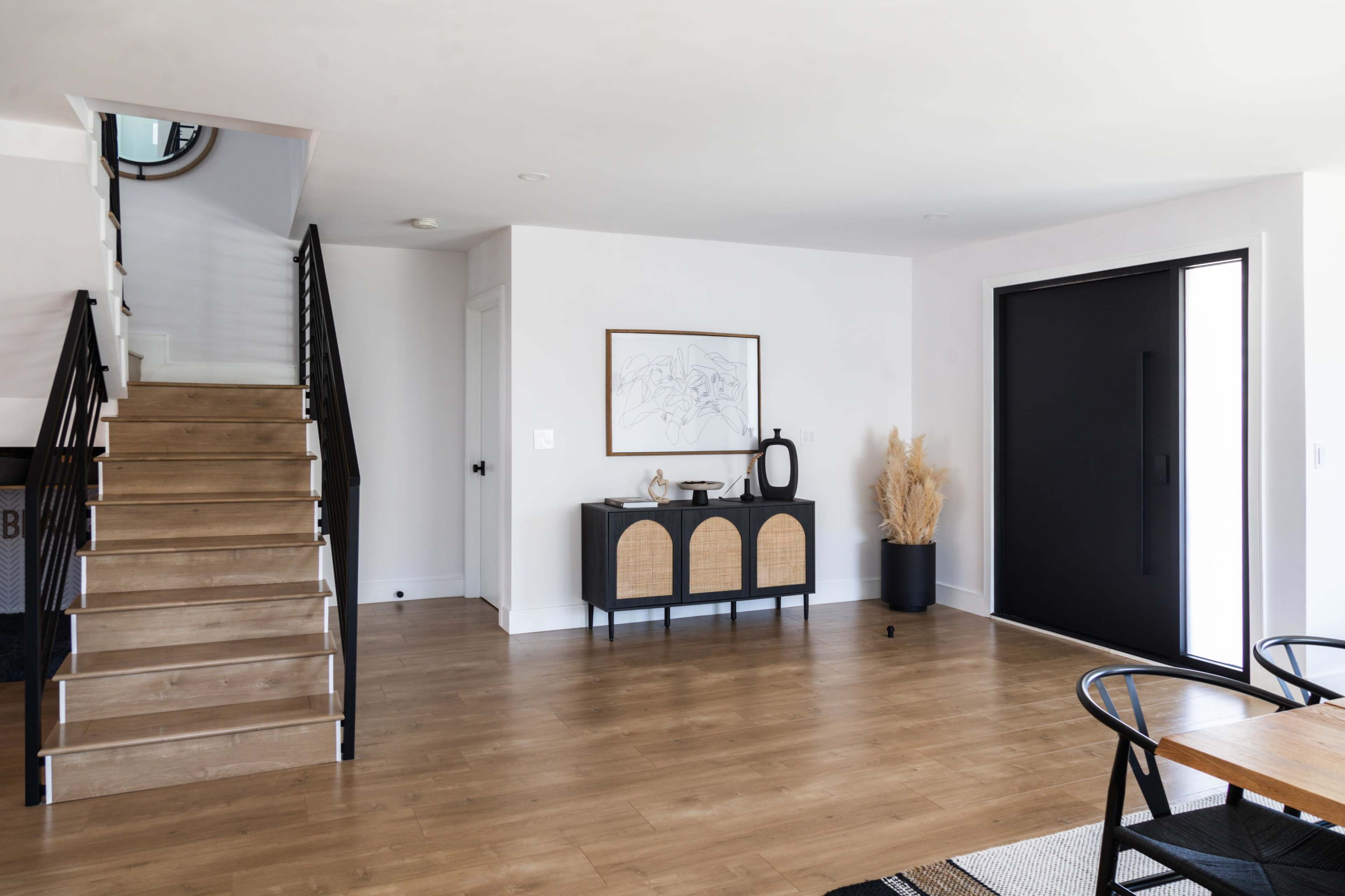 A modern entryway features a staircase on the left, a black front door, and a console table with decorative items against a light-colored wall.