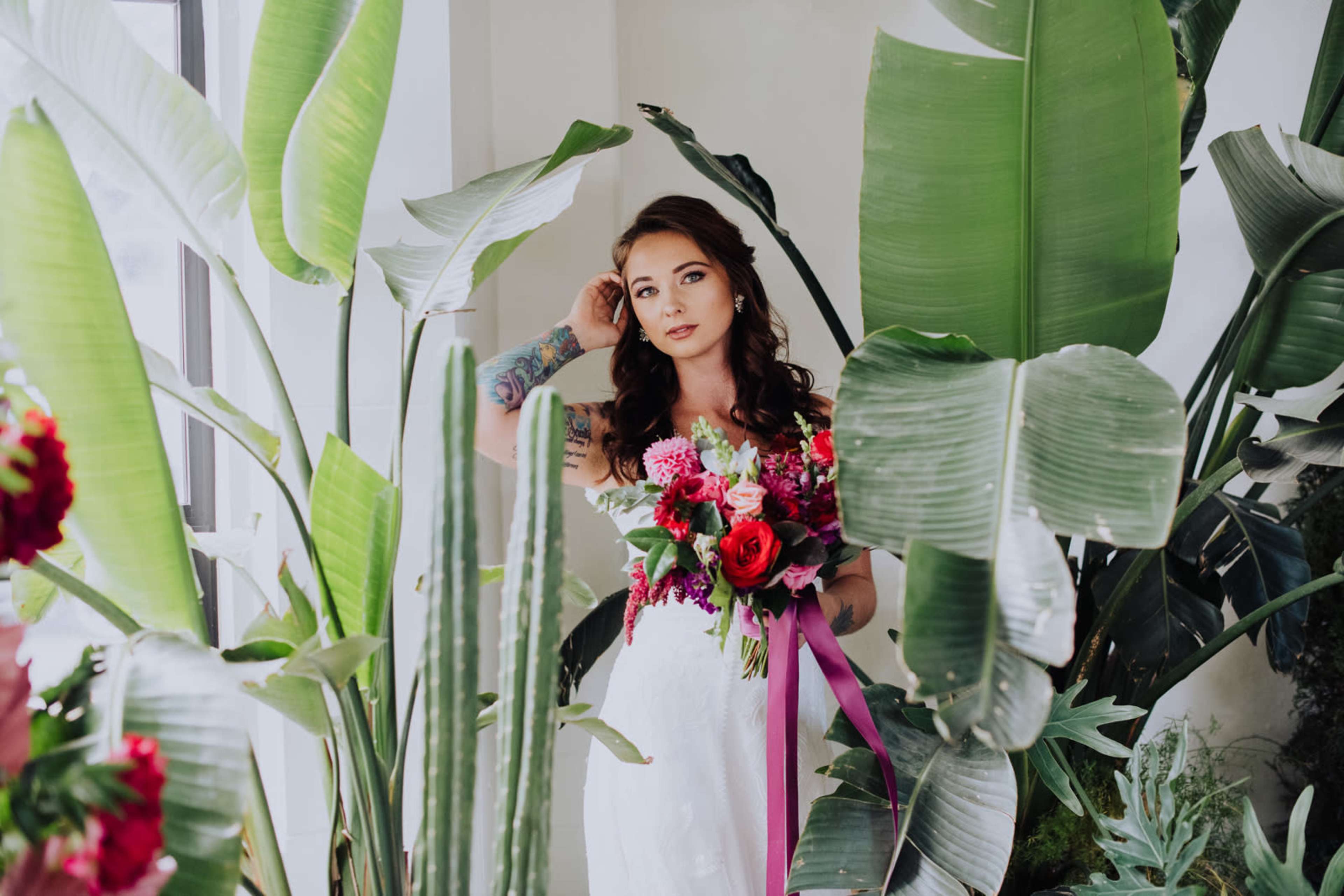A bride holds a bouquet of flowers while standing amidst large green plants in a bright room.