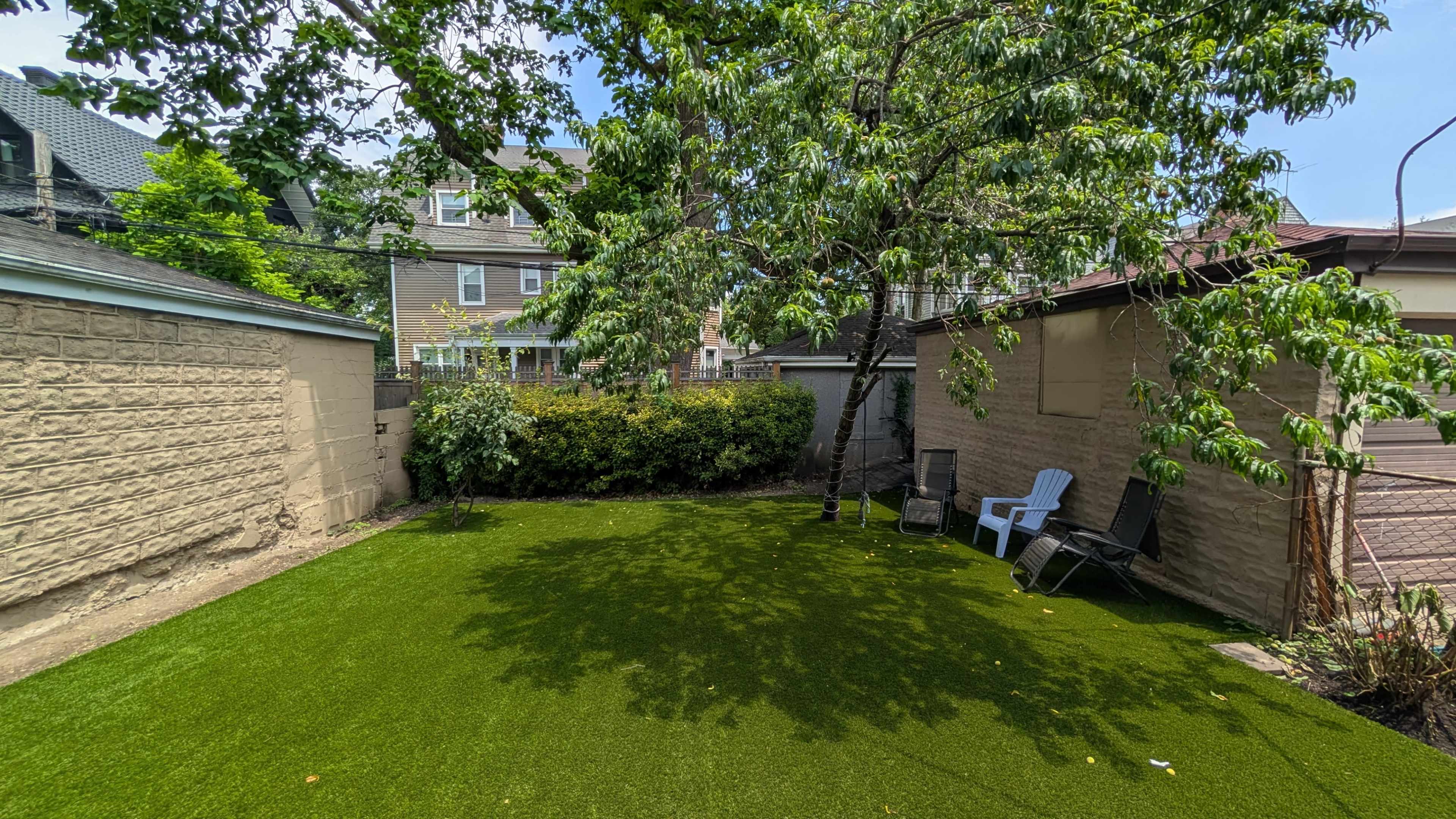 The image shows a small backyard with artificial turf, a tree providing shade, and a couple of lawn chairs near a wall and a garage.