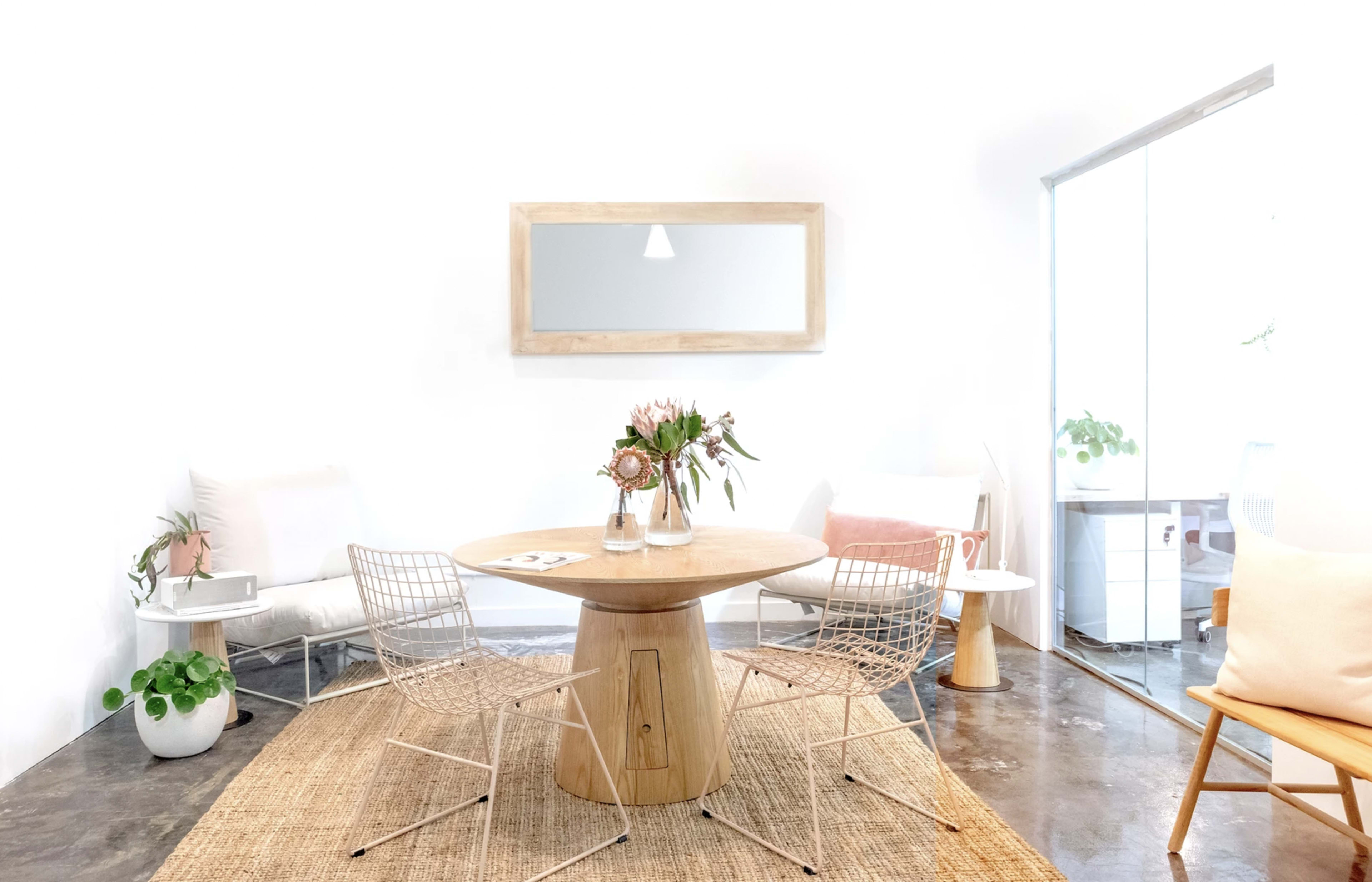 The image depicts a bright, modern interior featuring a round wooden table surrounded by wire chairs, with potted plants and a large mirror on a white wall.