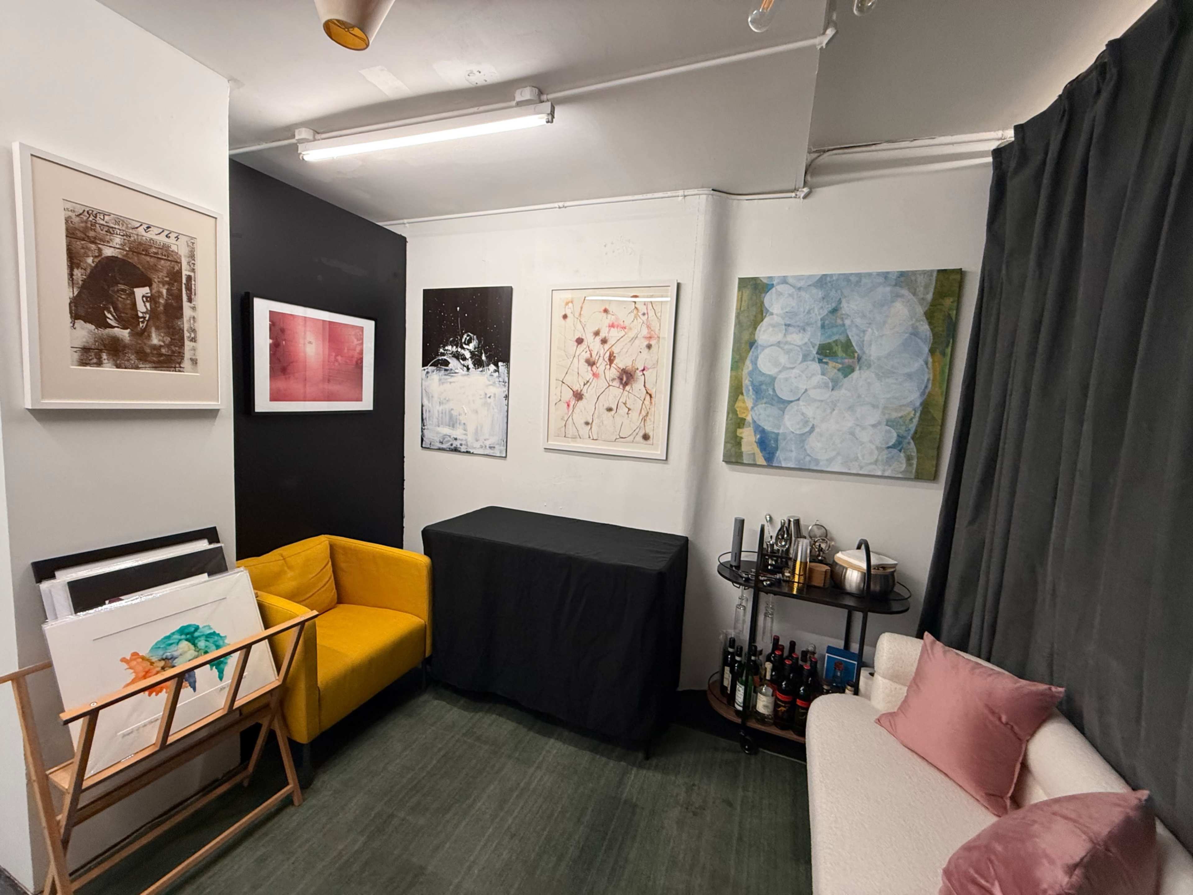 The image shows a small room featuring a yellow sofa, a white couch with pink cushions, several pieces of artwork on the walls, and a table with a black cloth and a bar cart.