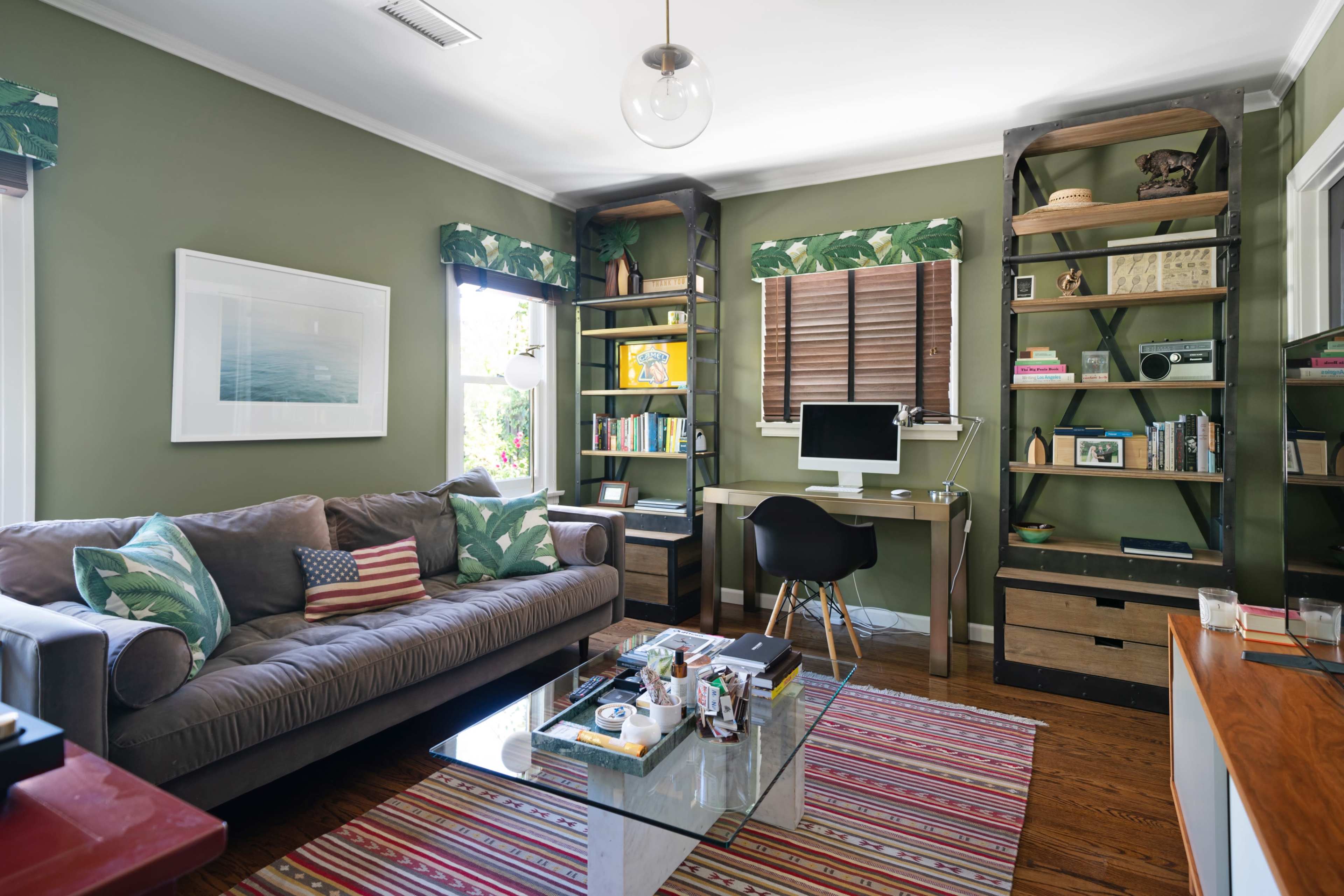A living room features a gray couch with decorative pillows, a striped rug, a glass coffee table, and a workspace with a computer against green walls adorned with shelves.