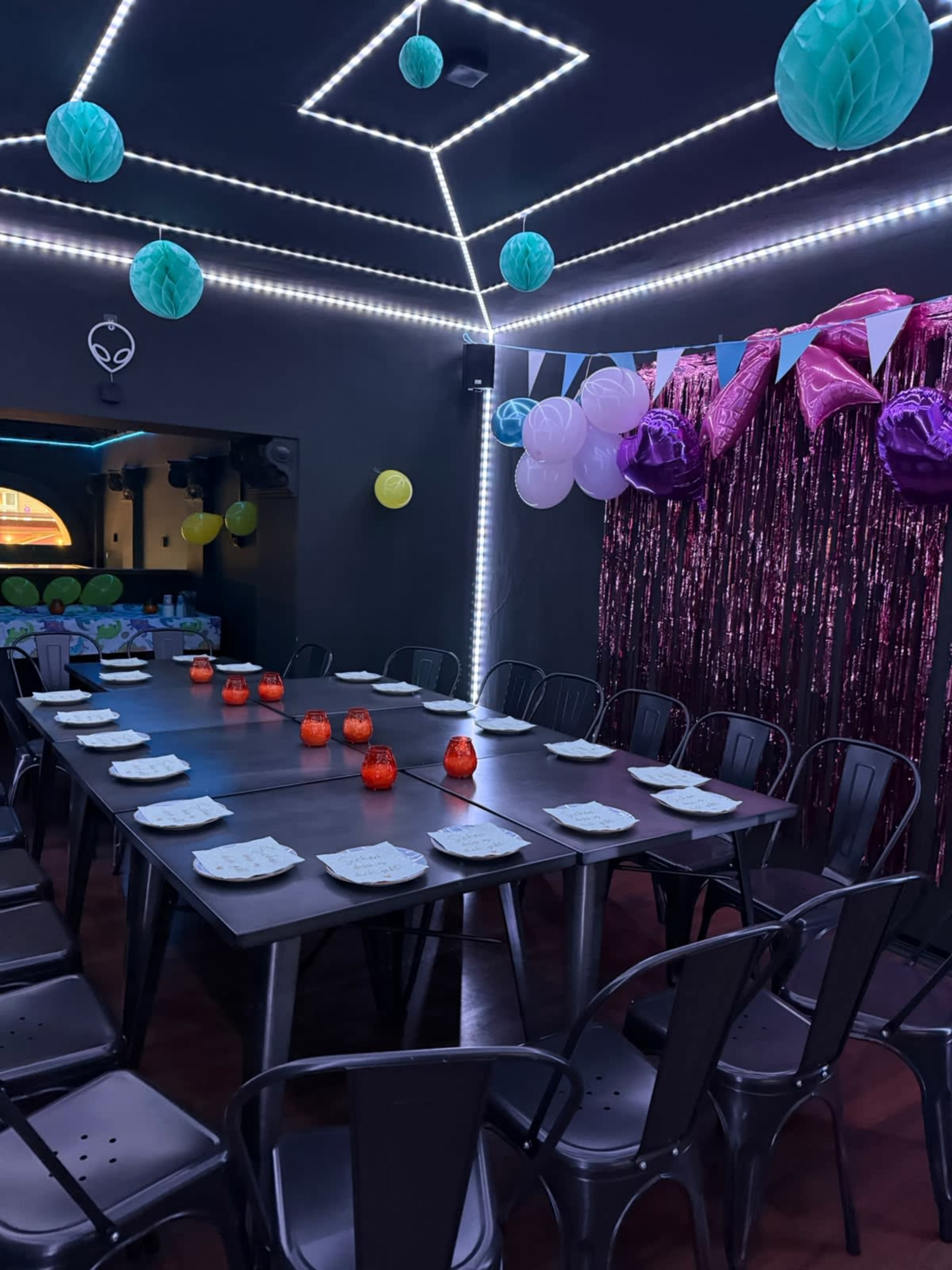 A long table set with plates and decorative candles is surrounded by black chairs in a room adorned with colorful balloons and lights.