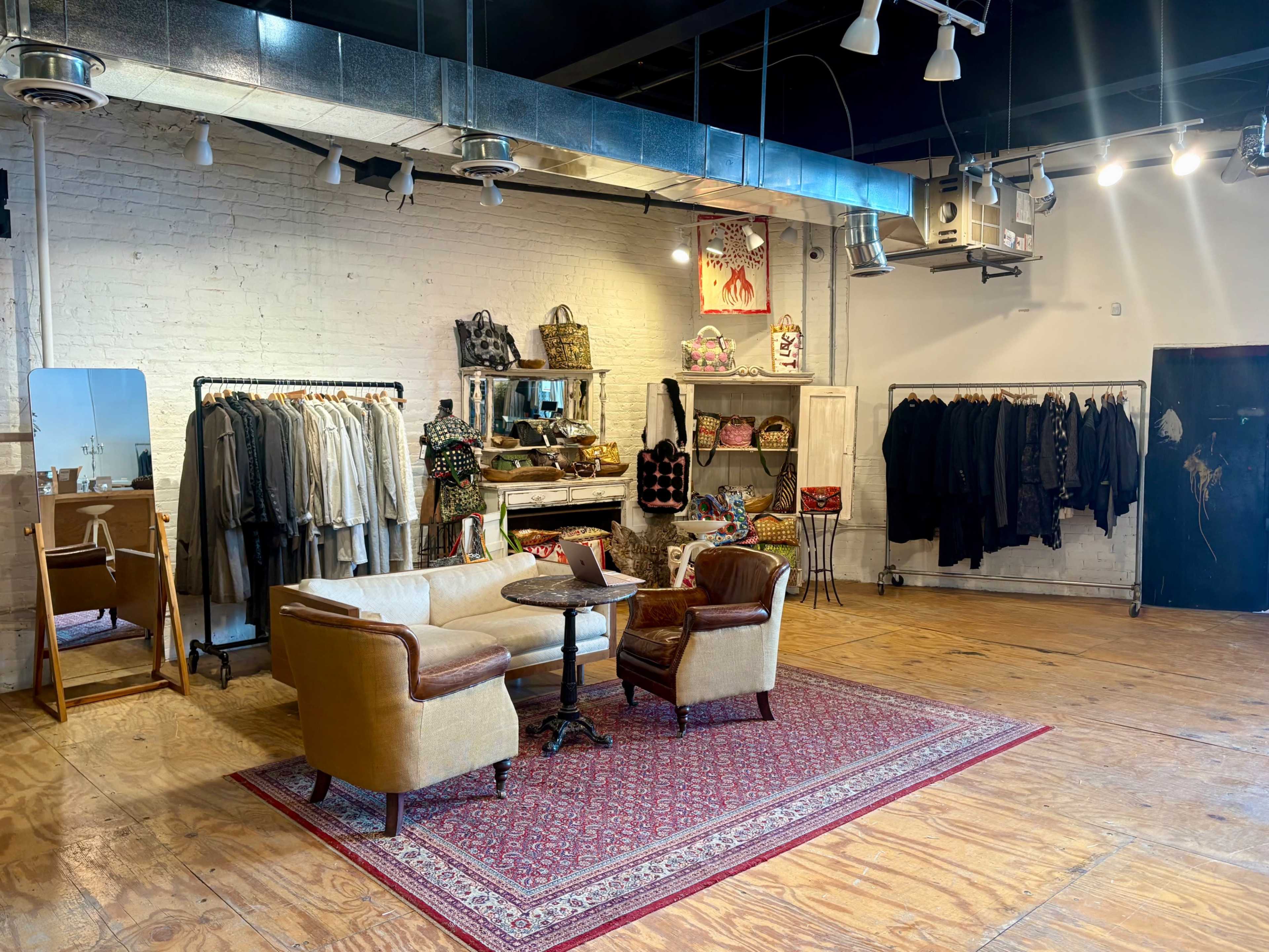 A clothing boutique features racks of garments, a mirror, and a seating area with two chairs arranged on a patterned rug.