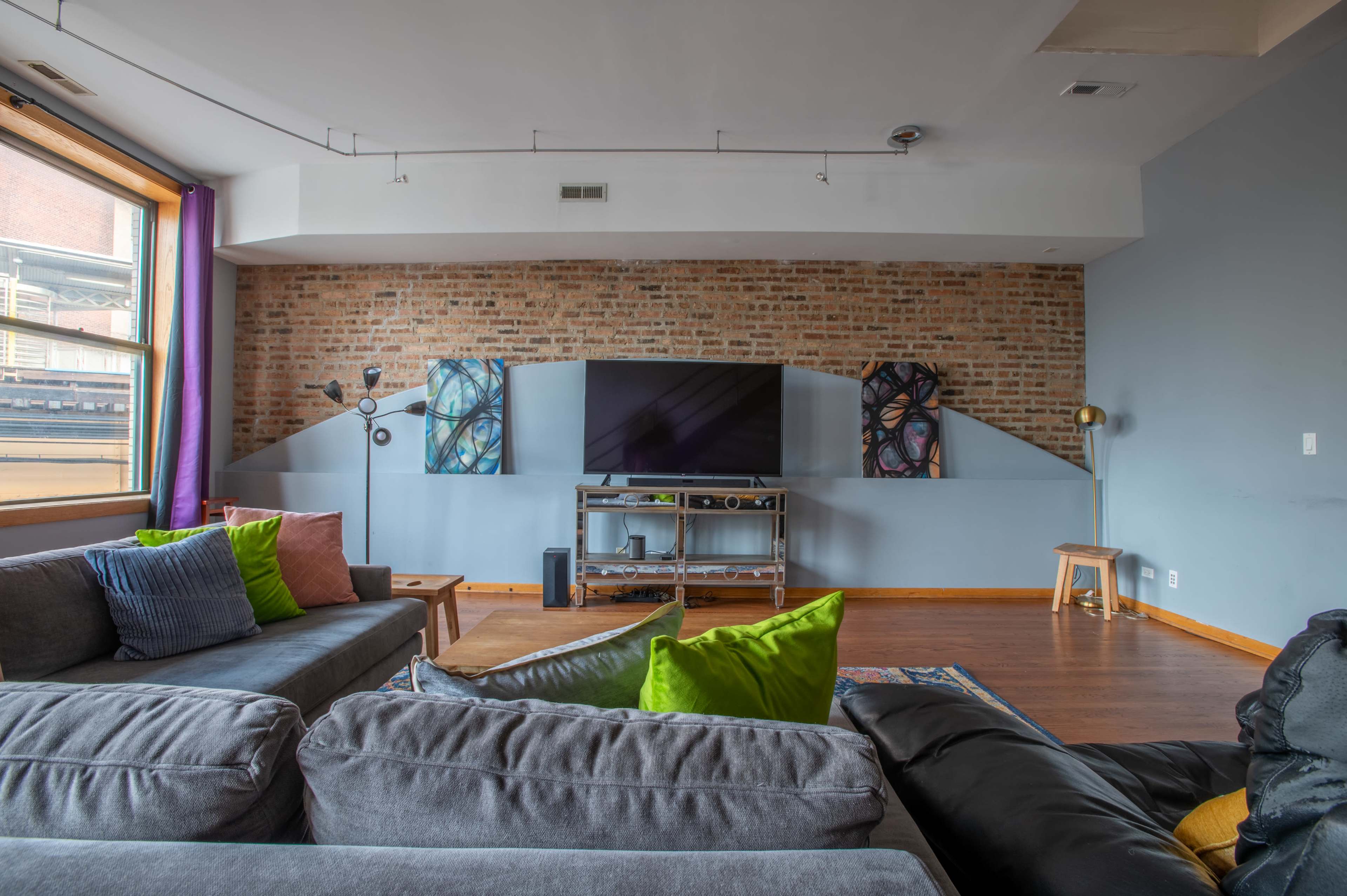 A modern living room features a sectional sofa, a wall-mounted television, and colorful artwork on exposed brick.