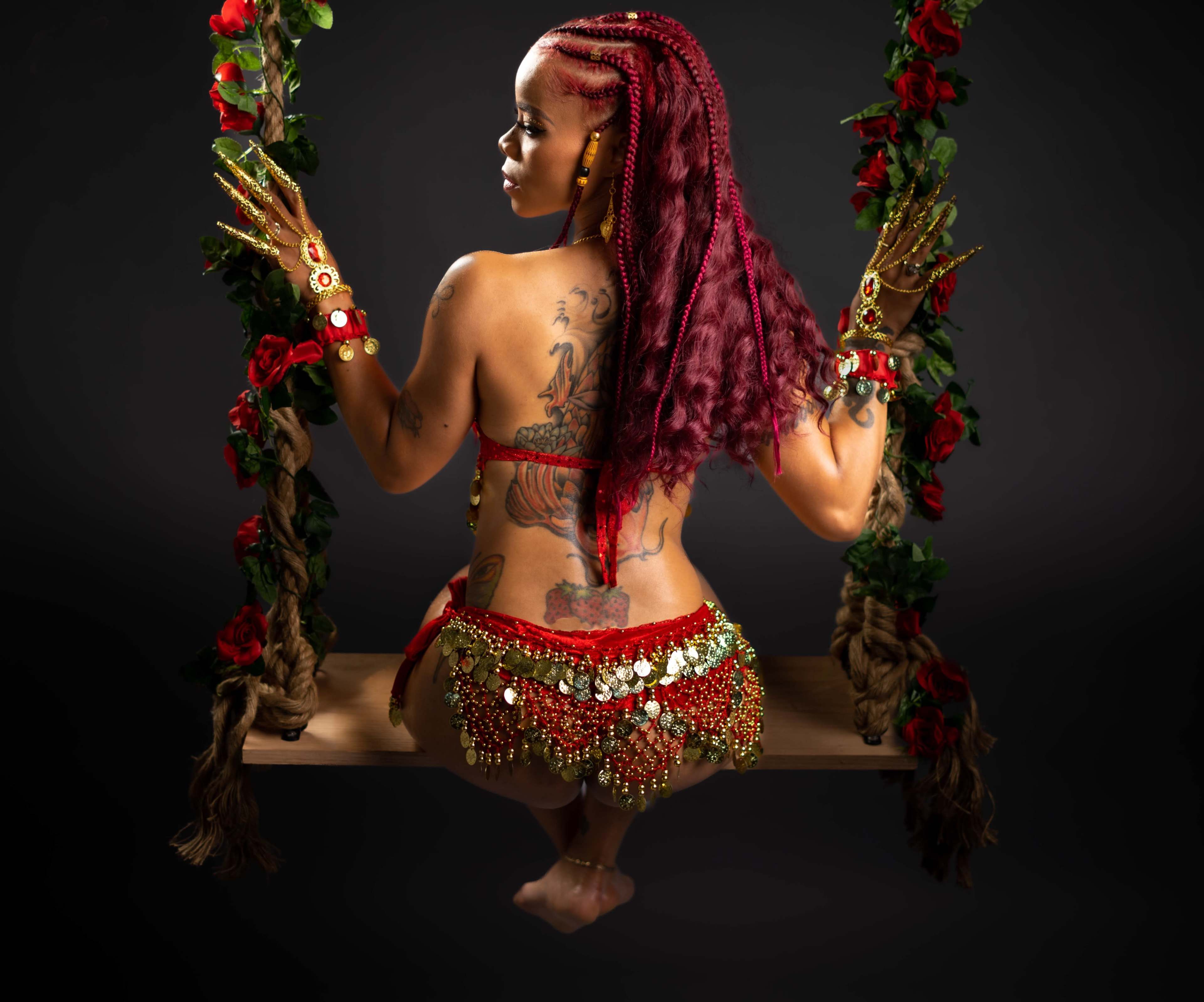 A woman with braided hair and intricate tattoos sits on a swing adorned with flowers, wearing a decorative outfit with sequins.