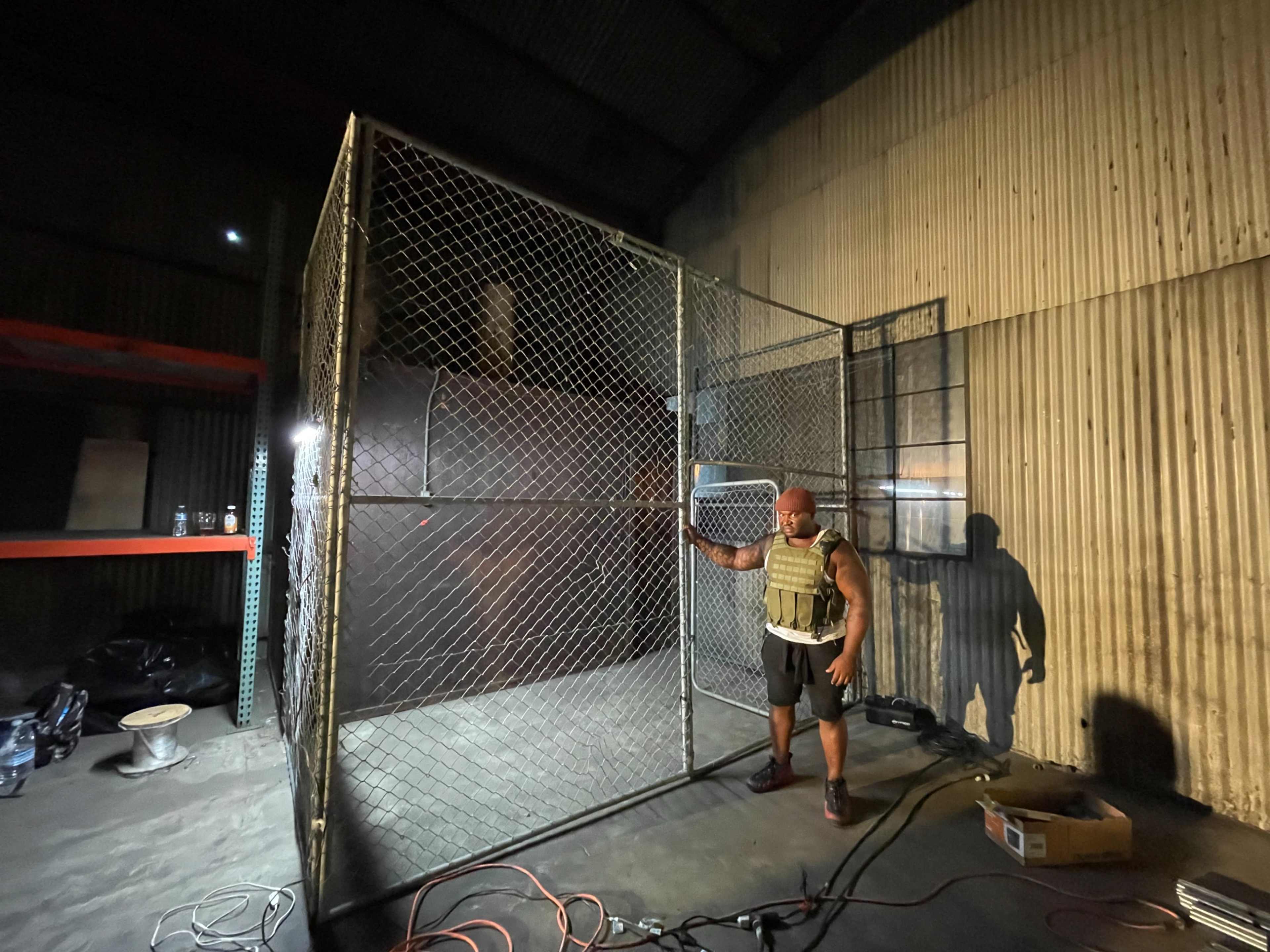 A man stands beside a large chain-link cage in a dimly lit warehouse.