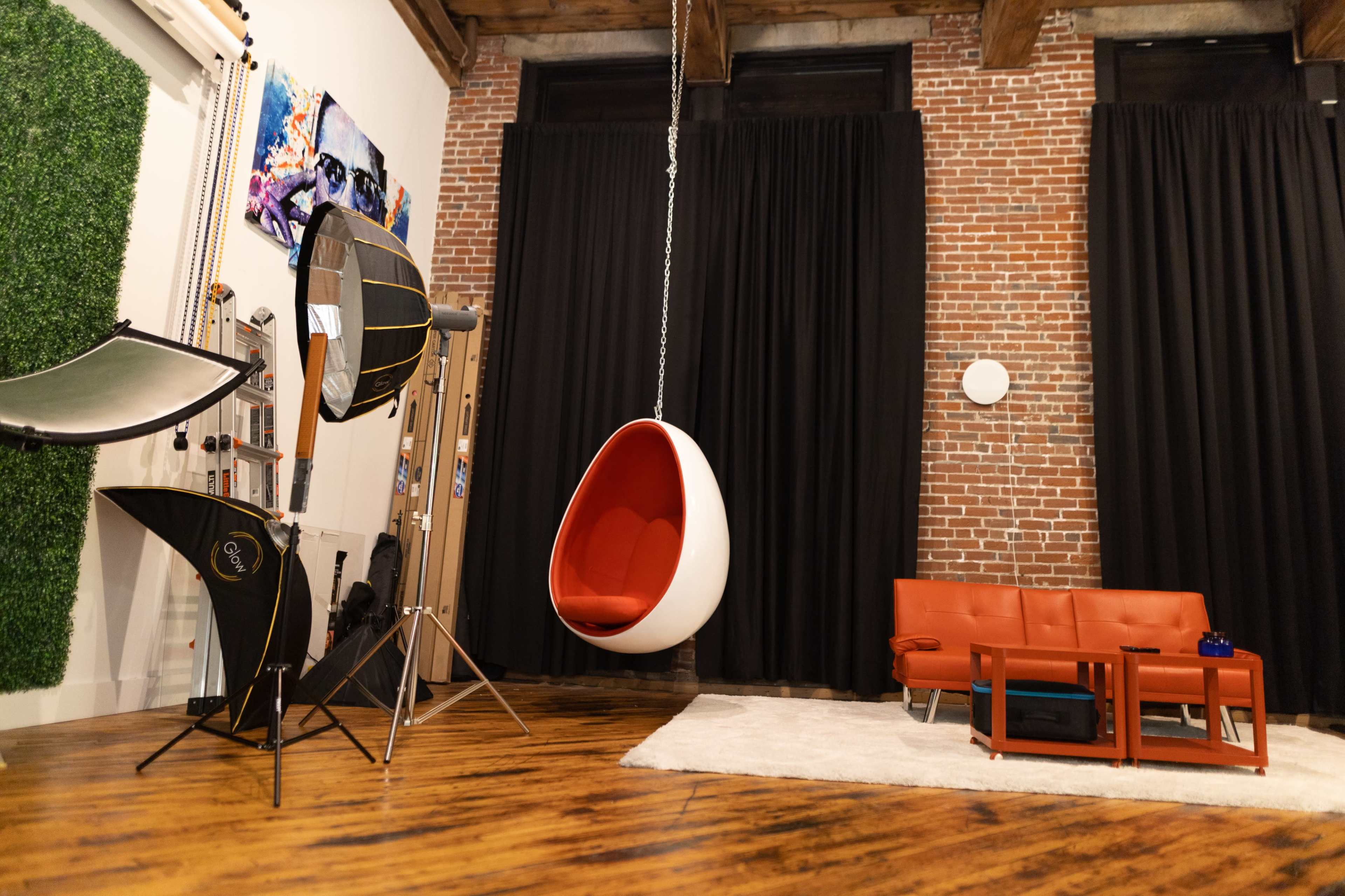 A modern studio space featuring a hanging egg chair, a red couch, and various photography equipment against a backdrop of exposed brick and dark curtains.