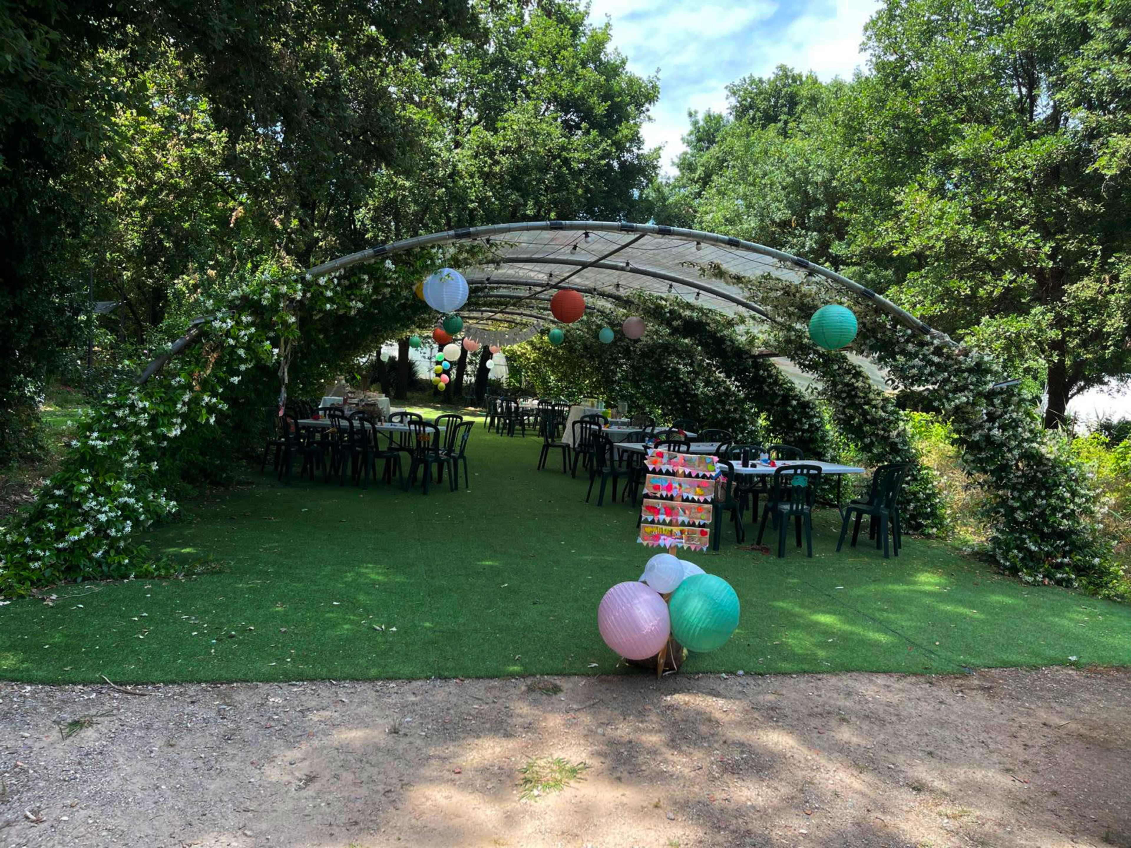 A tented area adorned with greenery and decorative lanterns is set up with tables and chairs on a grassy surface.