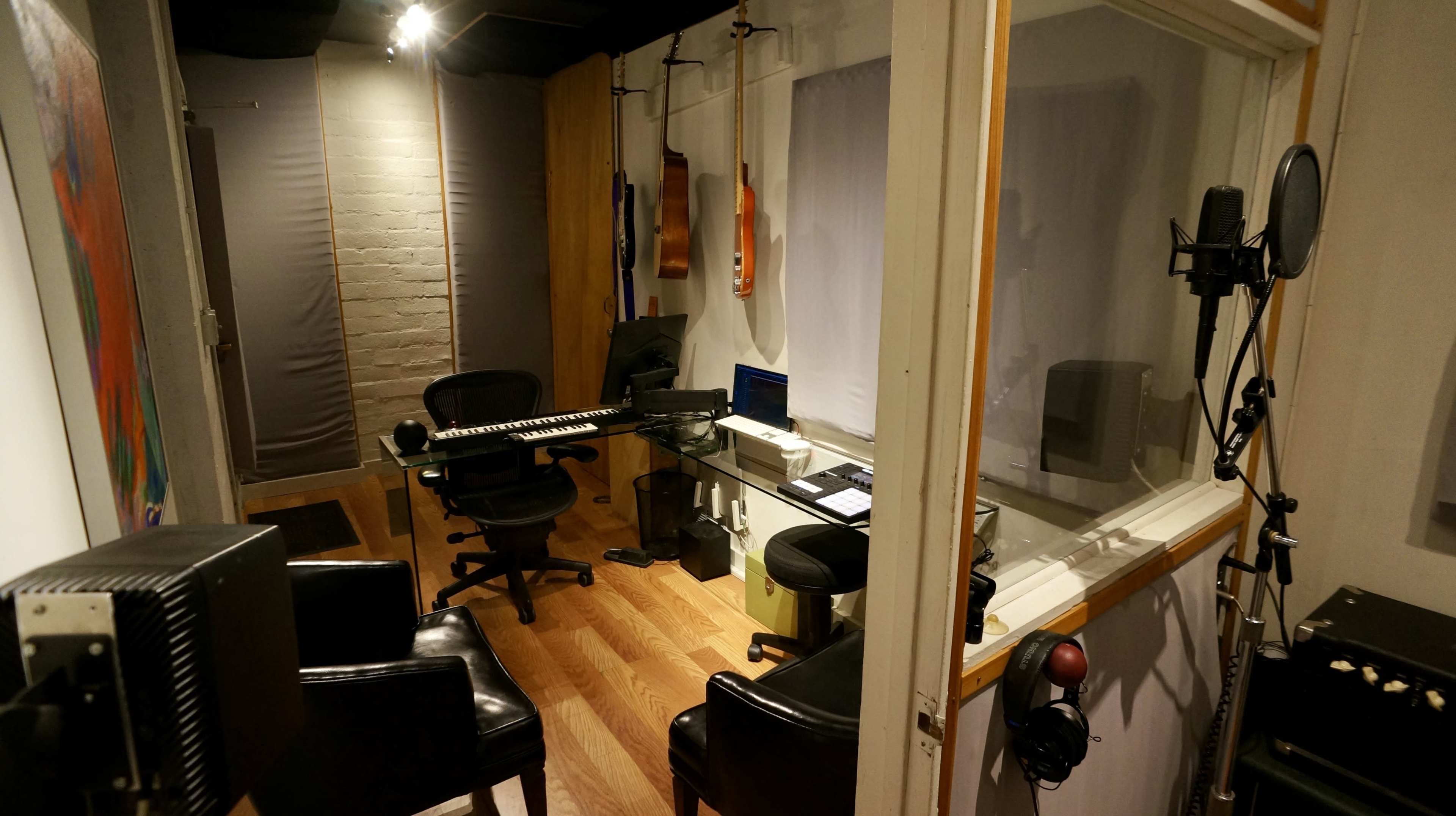 The image shows a small recording studio with a glass booth, a keyboard on a desk, and a few chairs.