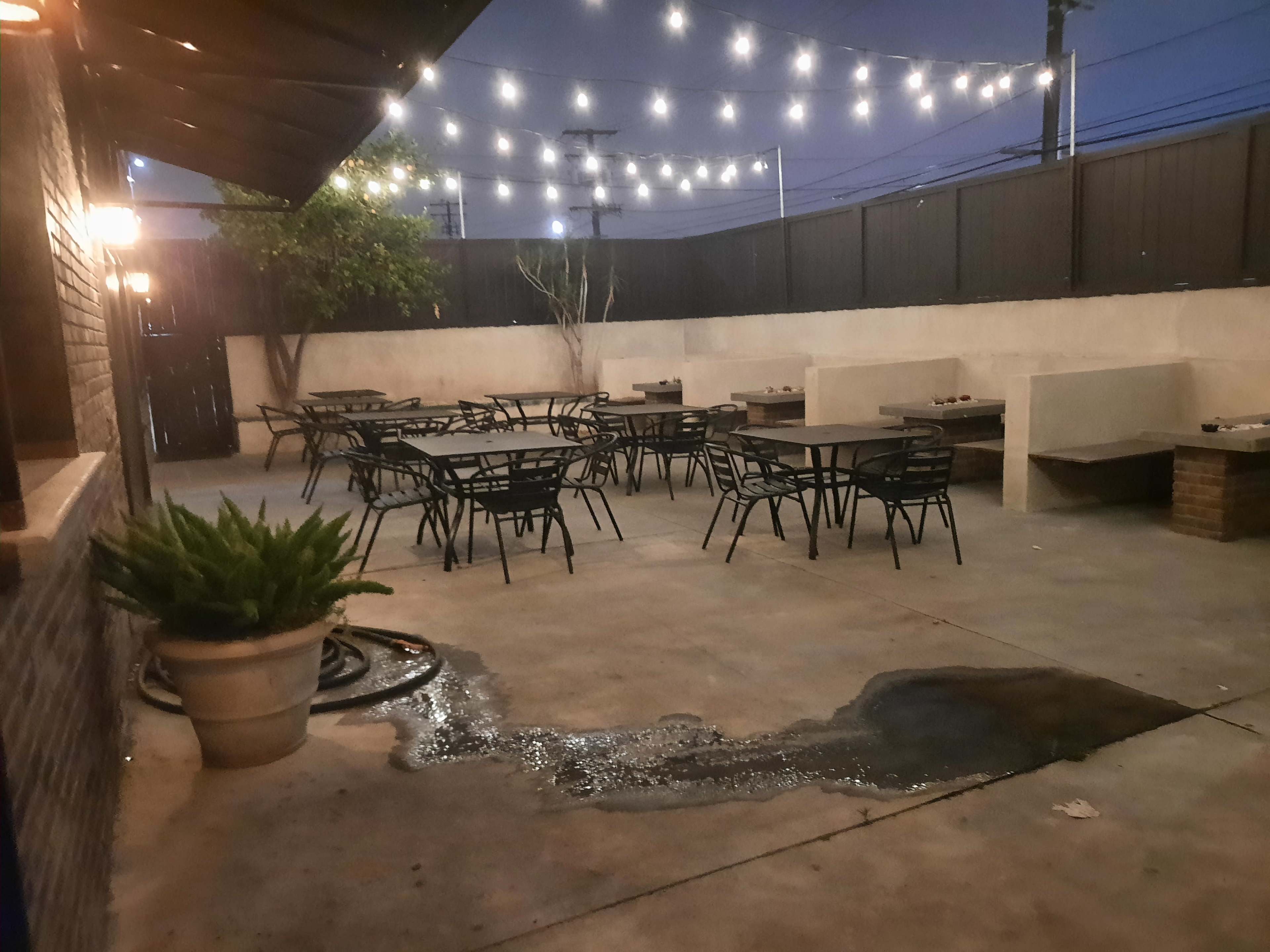An outdoor dining area features several tables and chairs under hanging lights, with a small puddle of water on the concrete floor.