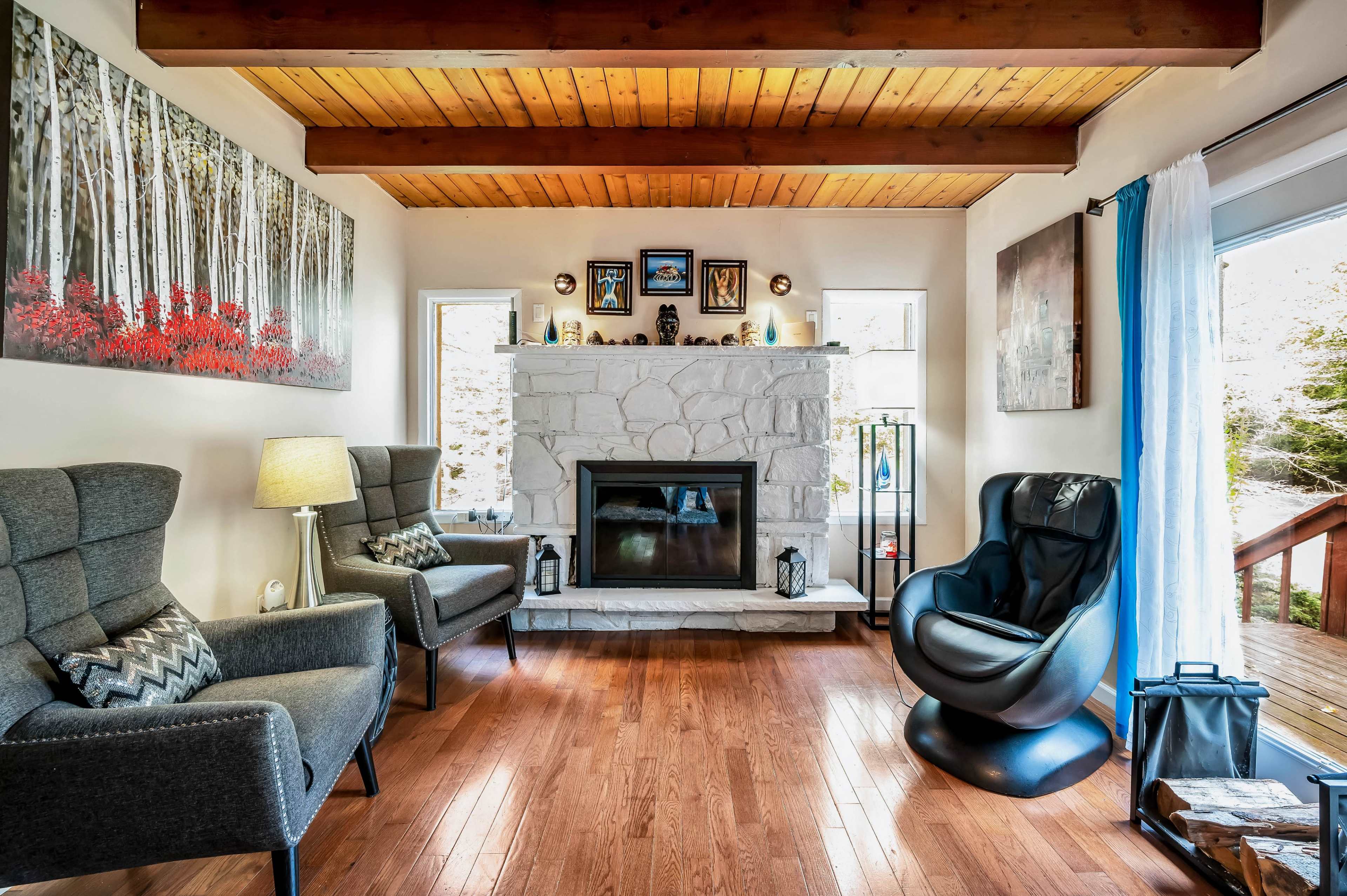 The living room features two armchairs and a modern black chair facing a stone fireplace, with wooden beams overhead and large windows providing natural light.
