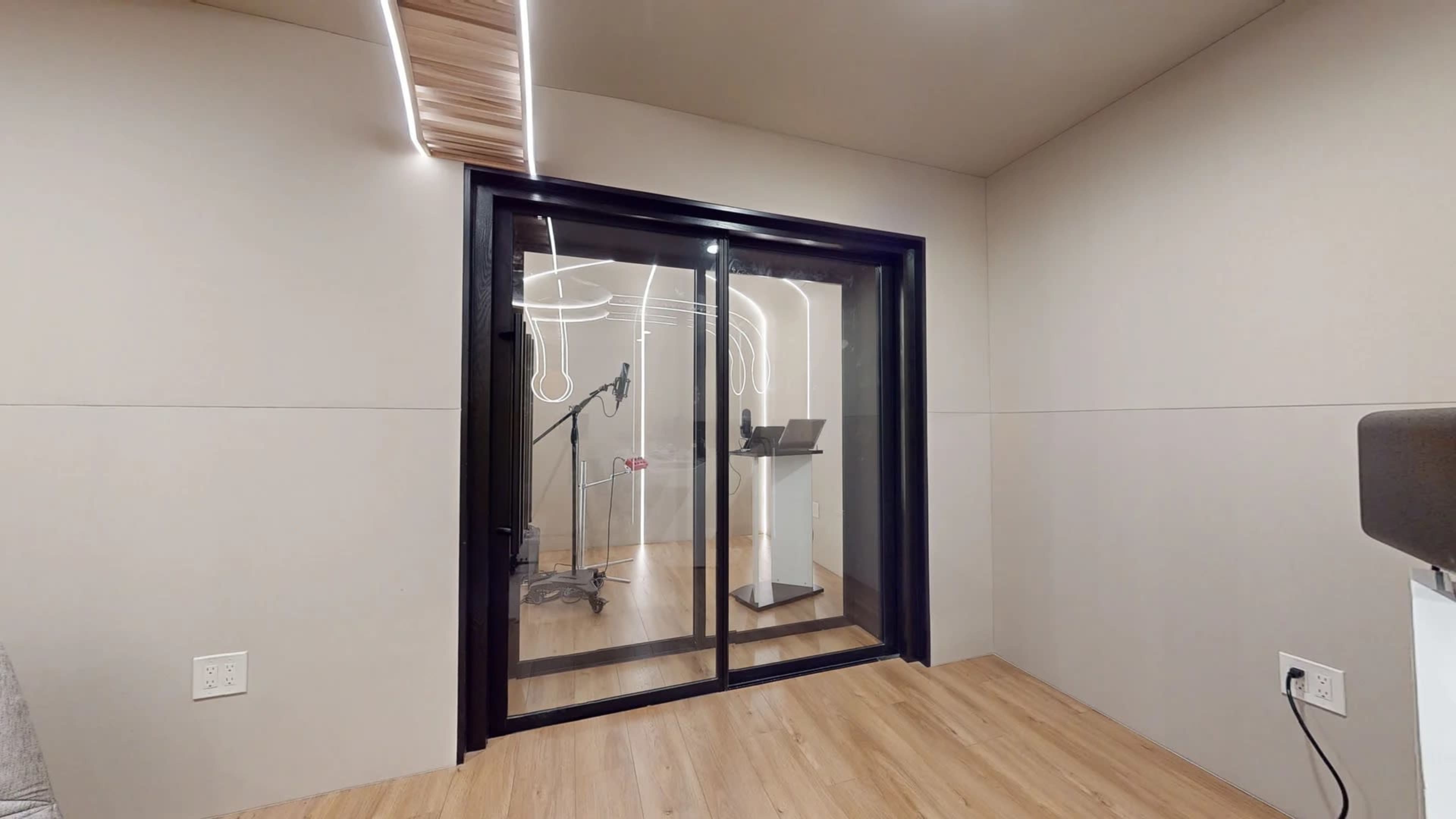 A small, minimalist recording booth with glass doors, a microphone setup, and wooden flooring.