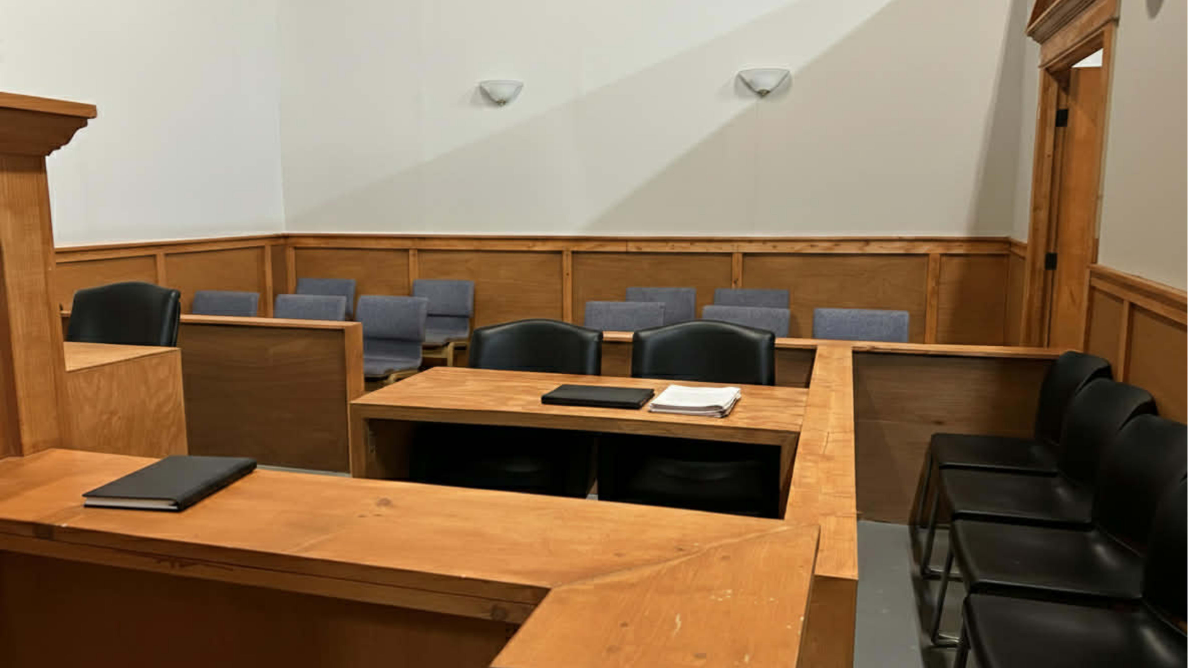 The image shows an empty courtroom with wooden paneling, a judge's bench, and rows of chairs for jurors and spectators.