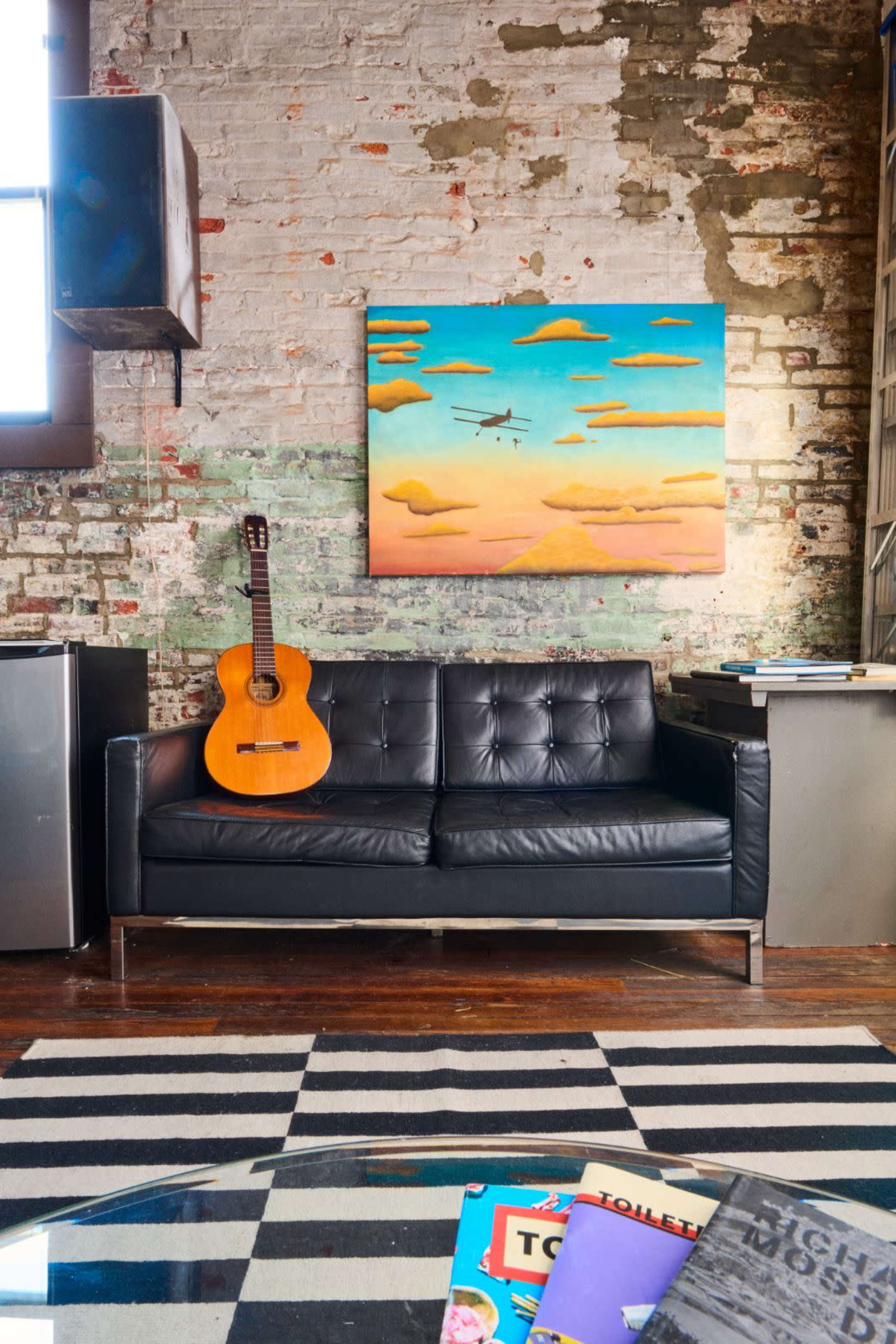 A black leather couch is positioned in front of a colorful painting of clouds on a brick wall, with a guitar resting against the couch and a striped rug beneath a glass coffee table displaying magazines.