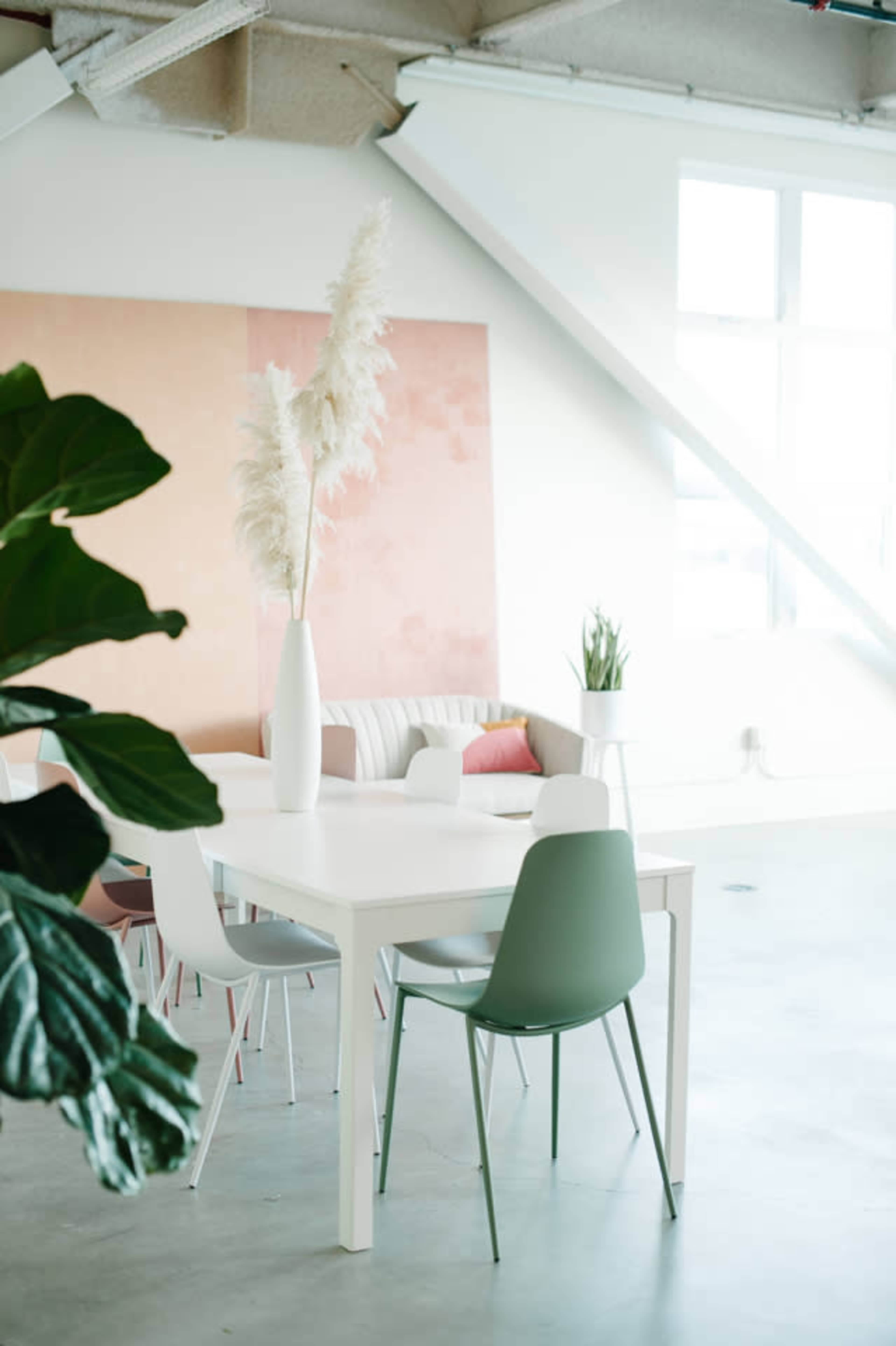 A bright, modern dining area features a white table surrounded by colorful chairs and a tall vase with pampas grass, set against a soft pink wall.