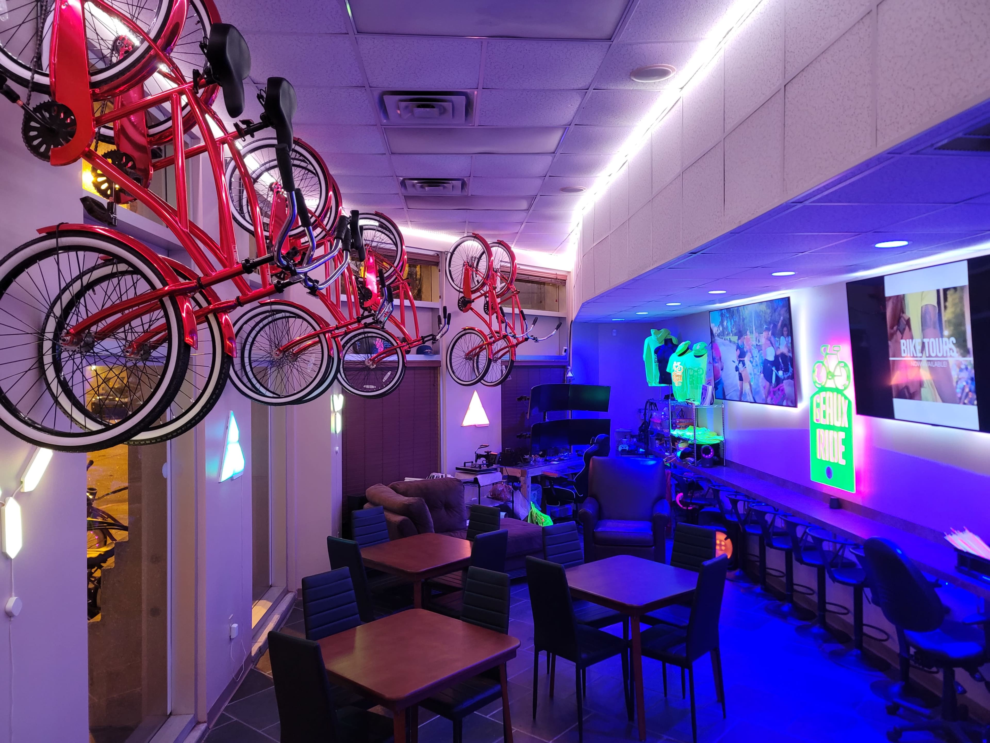 The interior of a vibrant café features hanging red bicycles, colorful neon lights, and a cozy seating area with dark wooden tables.