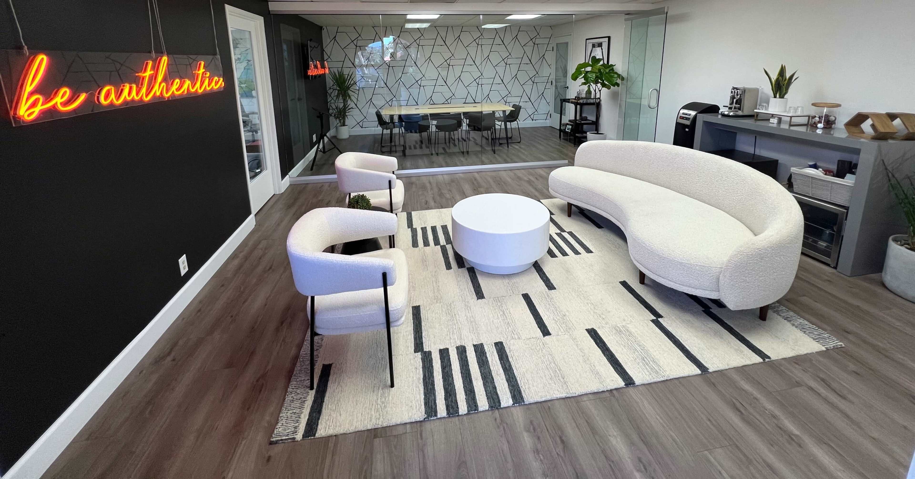 The image shows a modern office space with a seating area featuring a circular coffee table and a curved sofa, along with two armchairs on a patterned rug, and a glass-walled conference room in the background.