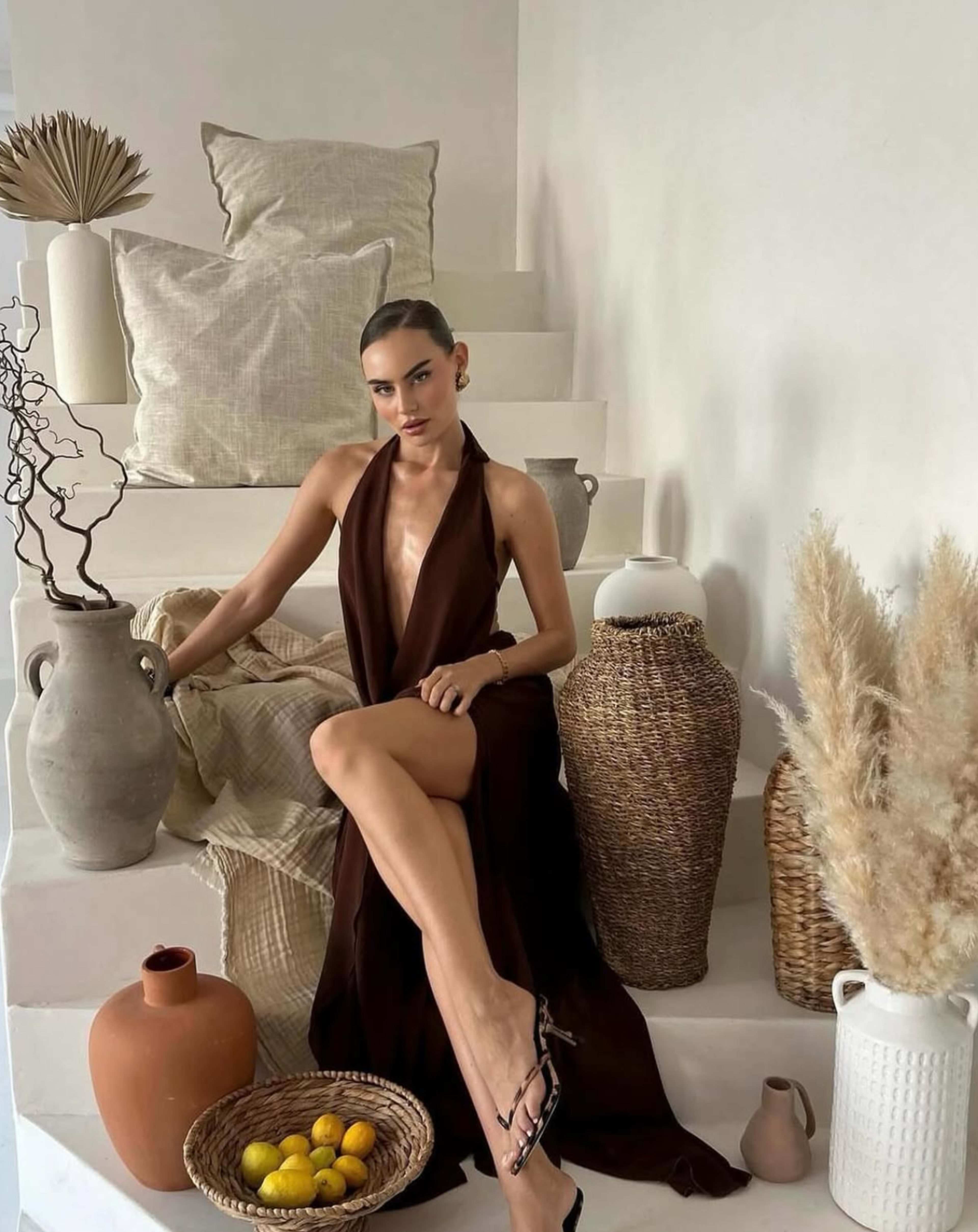 A woman in a brown dress sits on a staircase surrounded by decorative vases, baskets, and cushions.