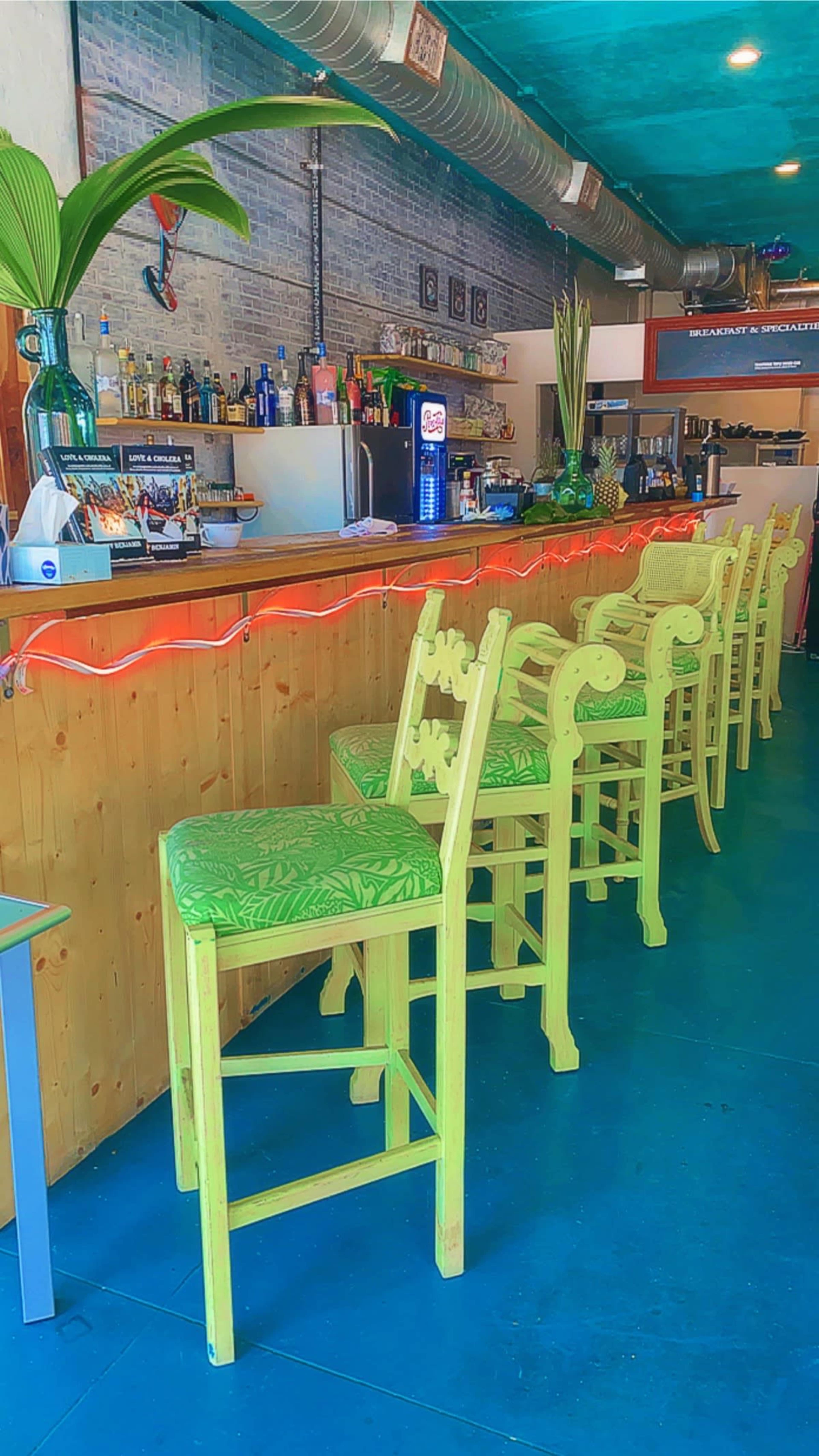 A brightly colored bar area with green stools featuring tropical-patterned cushions and a back counter displaying various bottles and beverages.