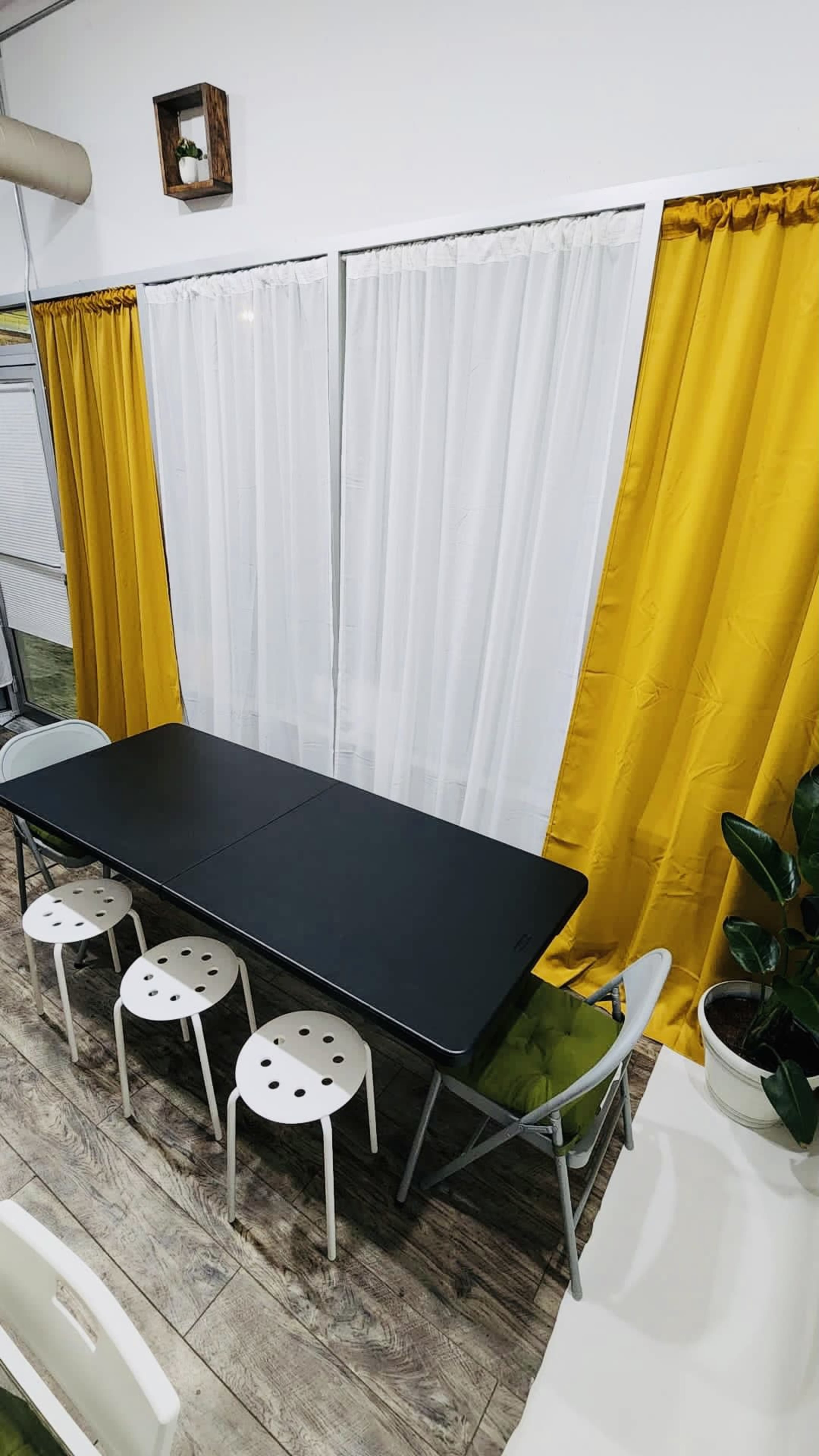 A black dining table is surrounded by four white stools in a room with yellow and white curtains.