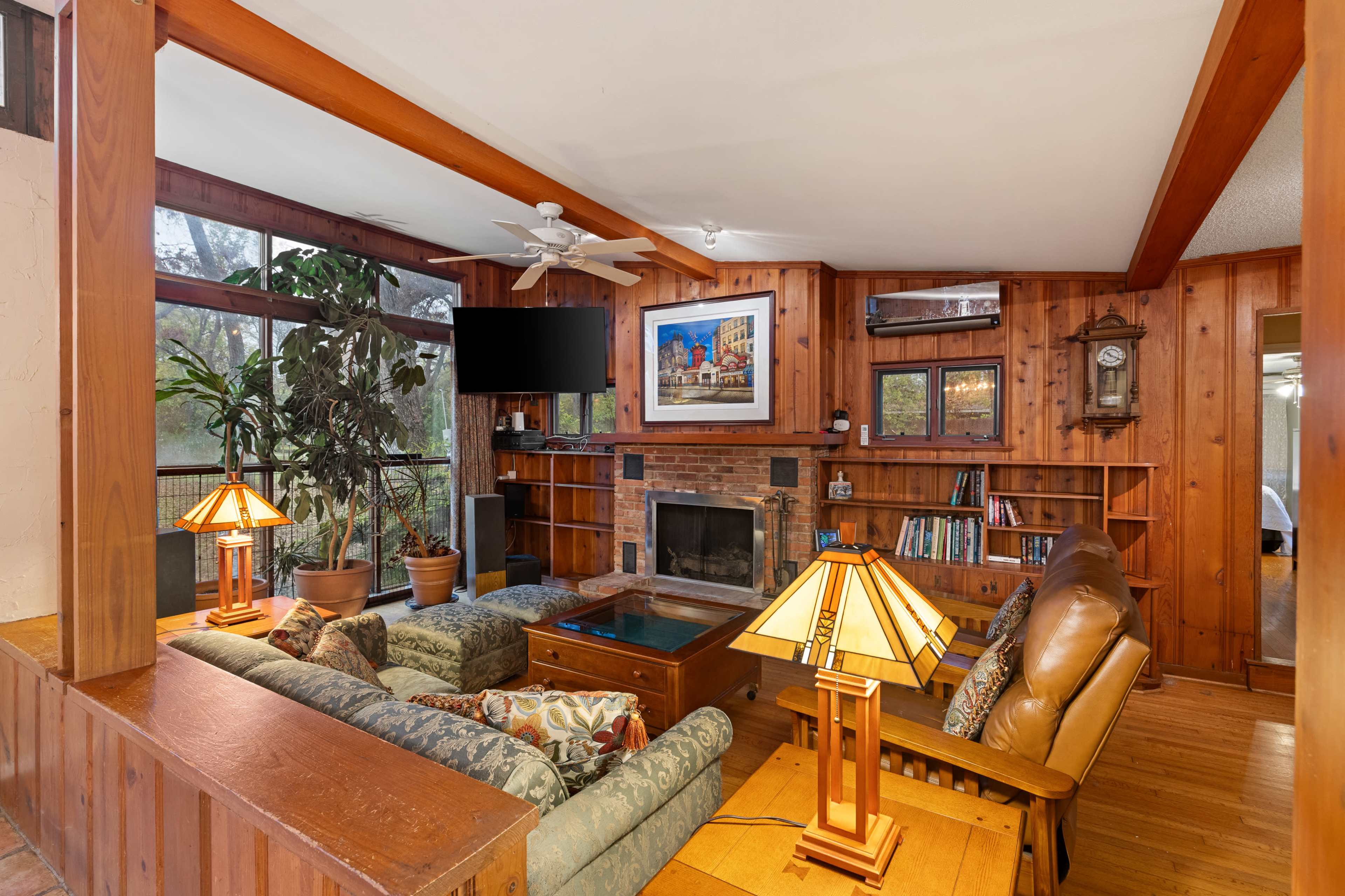 The image shows a cozy living room with wooden paneling, a brick fireplace, comfortable seating, and a large window overlooking greenery.