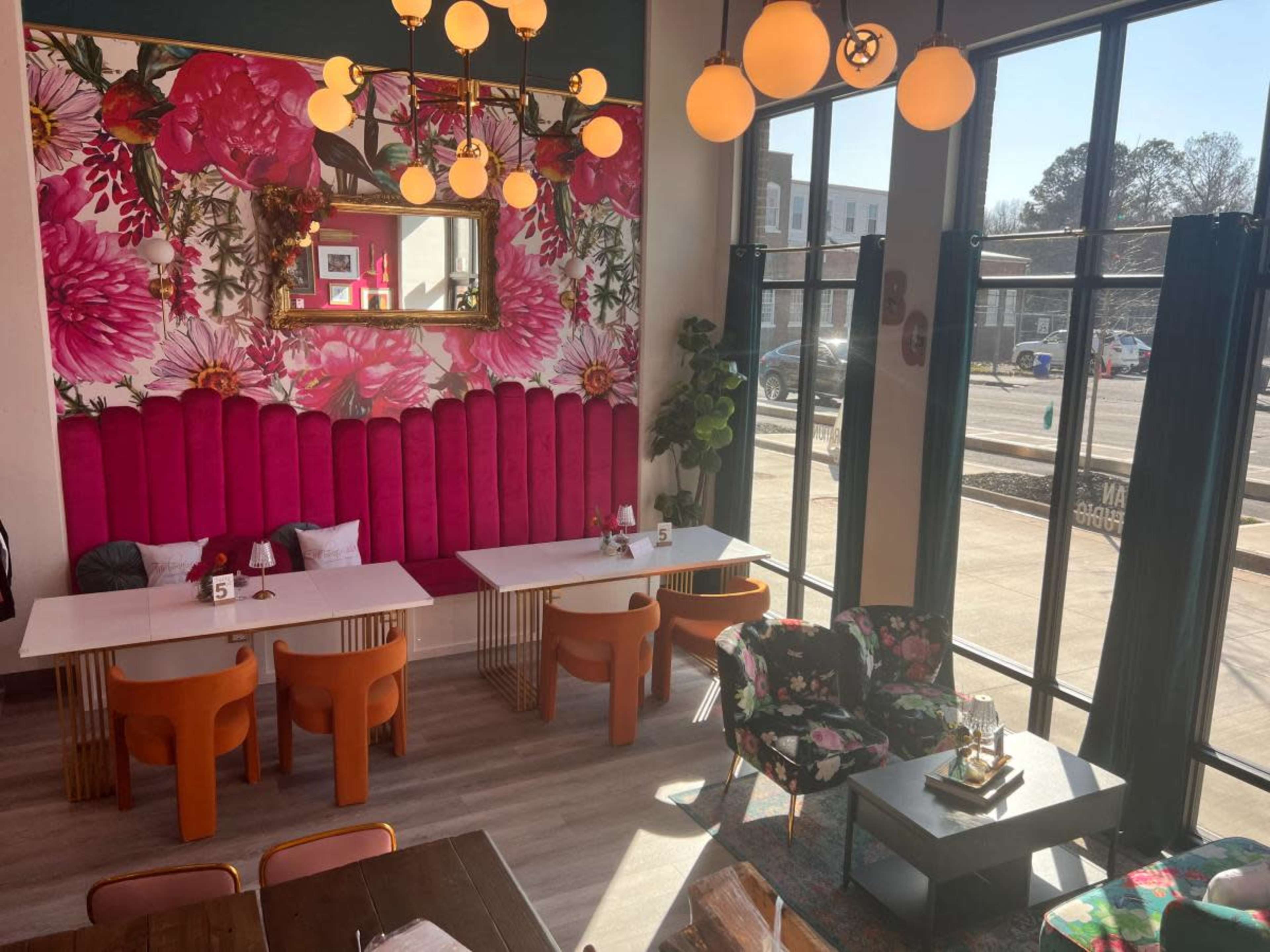 The image shows a modern café interior featuring a vibrant floral wallpaper, pink upholstered seating, and various tables with orange and floral-patterned chairs.