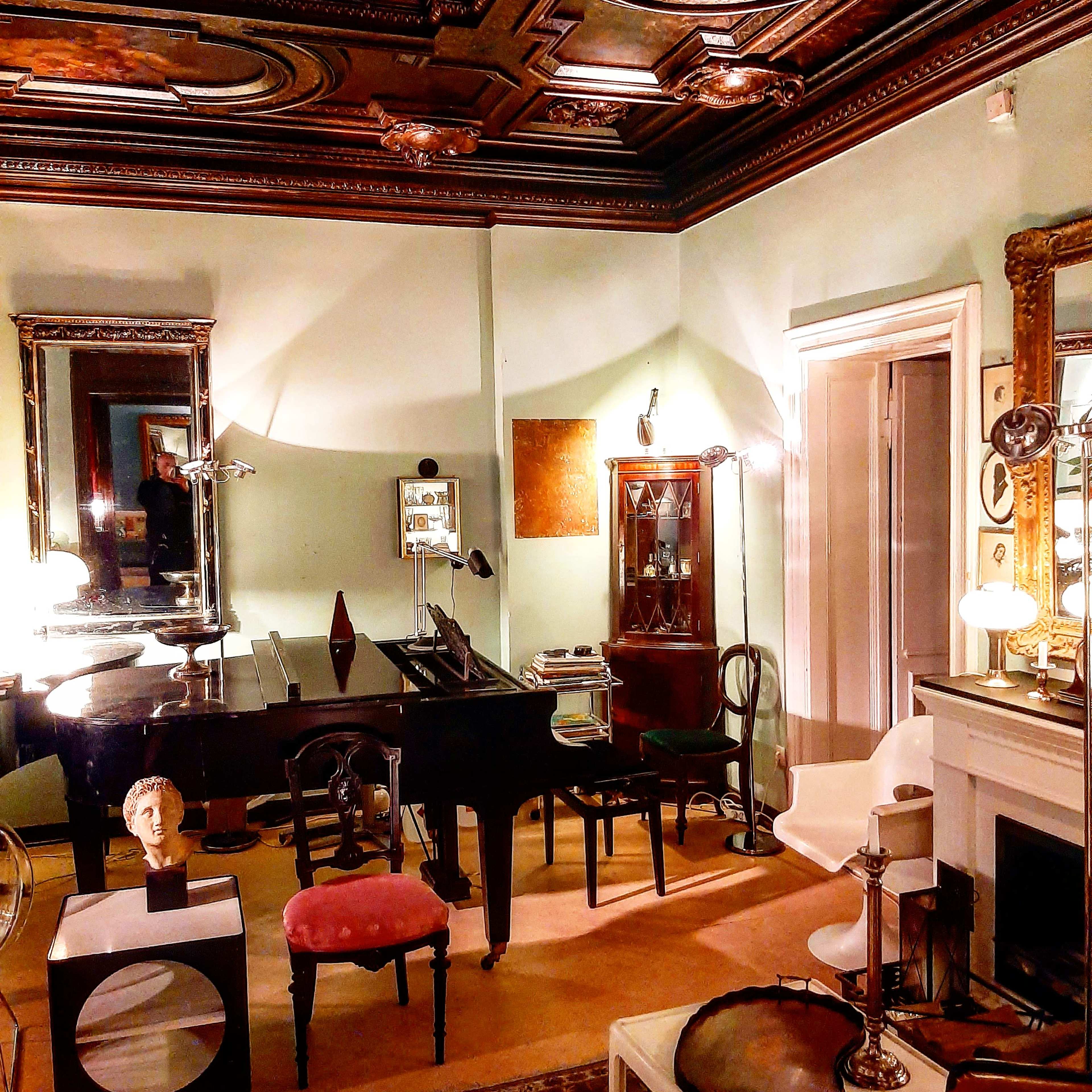 A vintage-style room featuring a grand piano, several ornate mirrors, a wooden cabinet, and various pieces of furniture arranged around a central area.