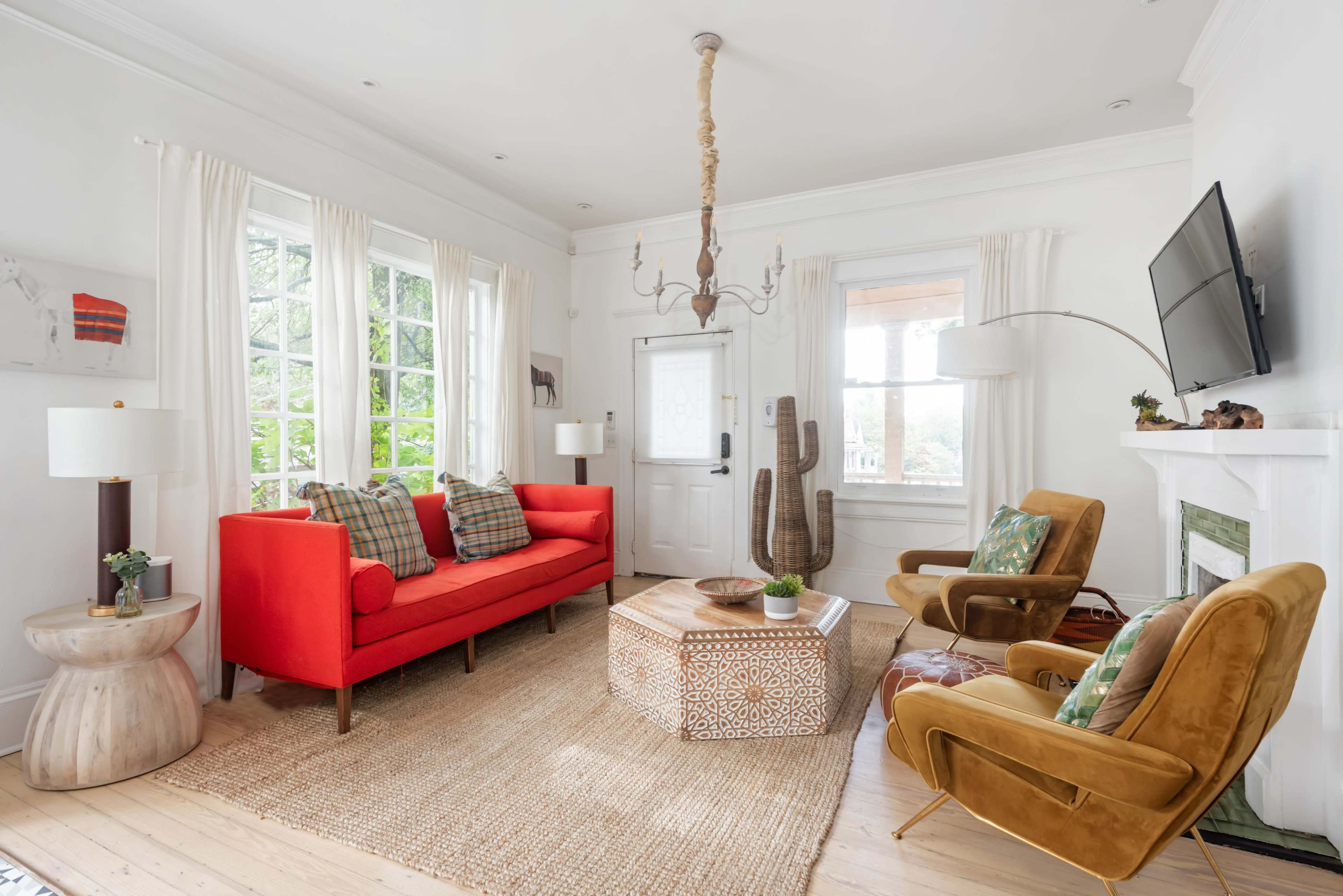 The image shows a bright living room with a red couch, brown armchairs, a patterned coffee table, and large windows letting in natural light.