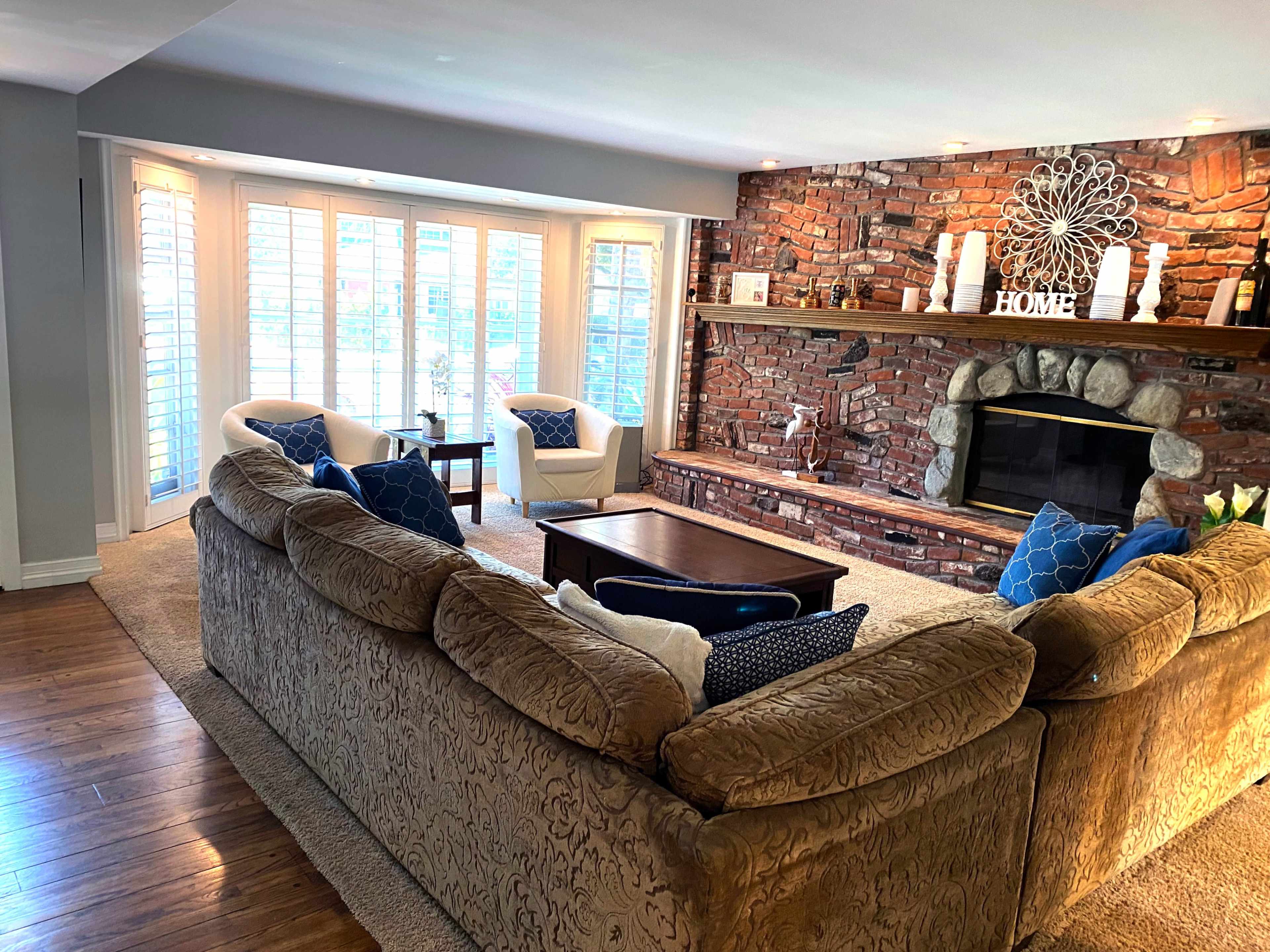 A spacious living room features a large sectional sofa, two armchairs, a coffee table, and a brick fireplace with decorative items on the mantel.