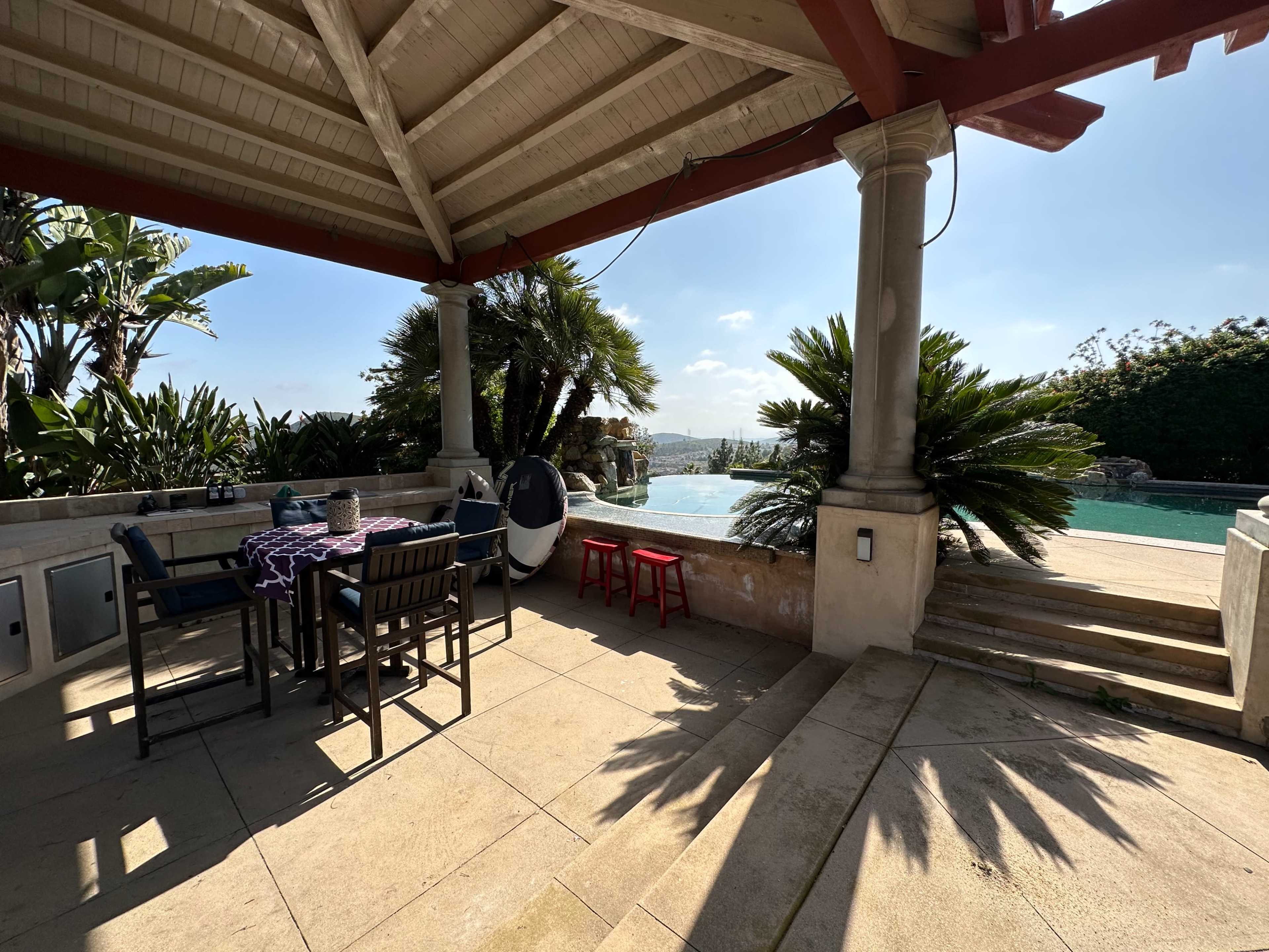 The image shows a outdoor seating area beside a pool, featuring a table and chairs under a covered porch with tropical plants in the background.