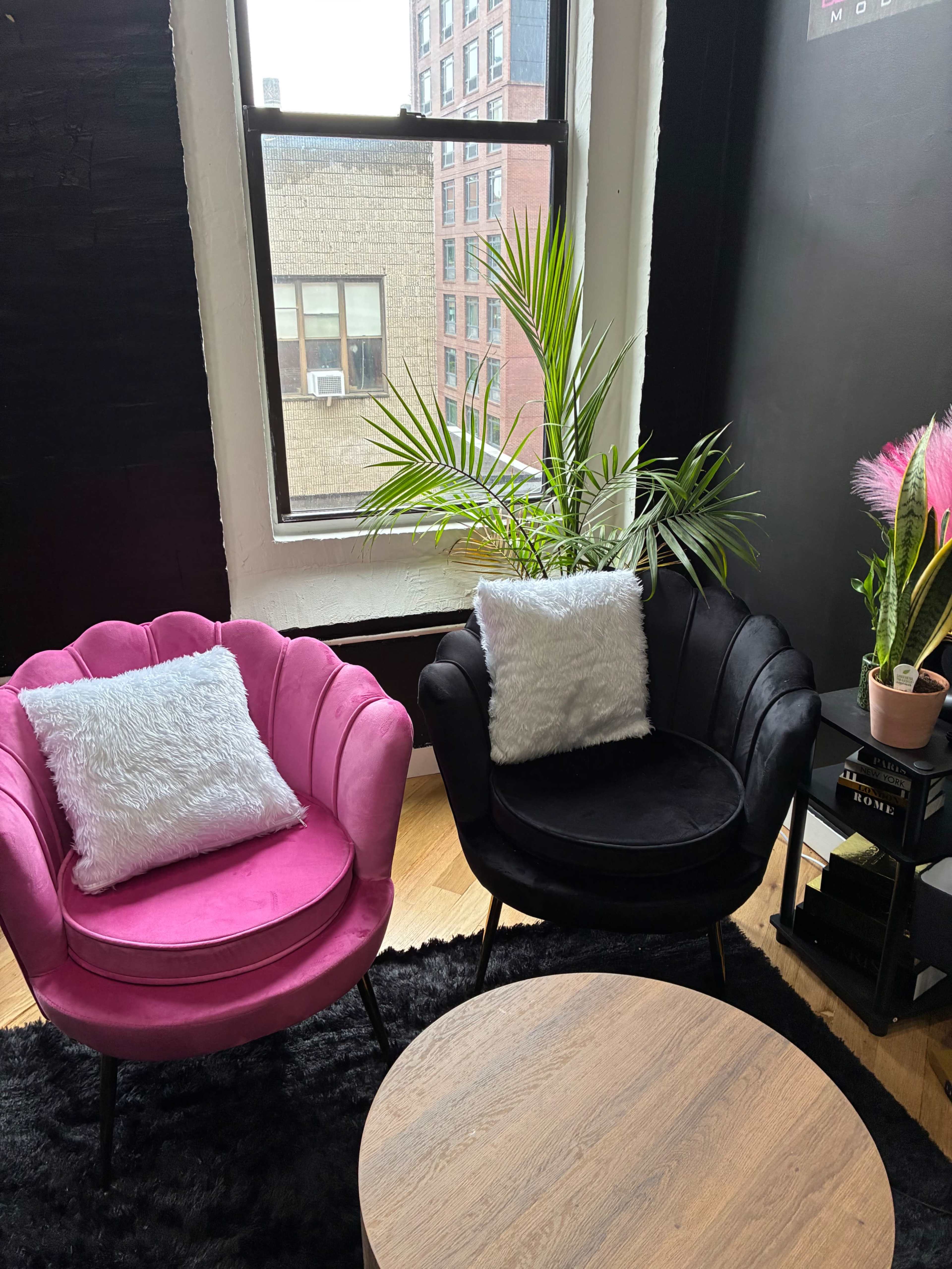 The image shows two stylish chairs, one pink and one black, with white pillows, positioned near a window with a view of a building, accompanied by a small round wooden coffee table and some indoor plants.