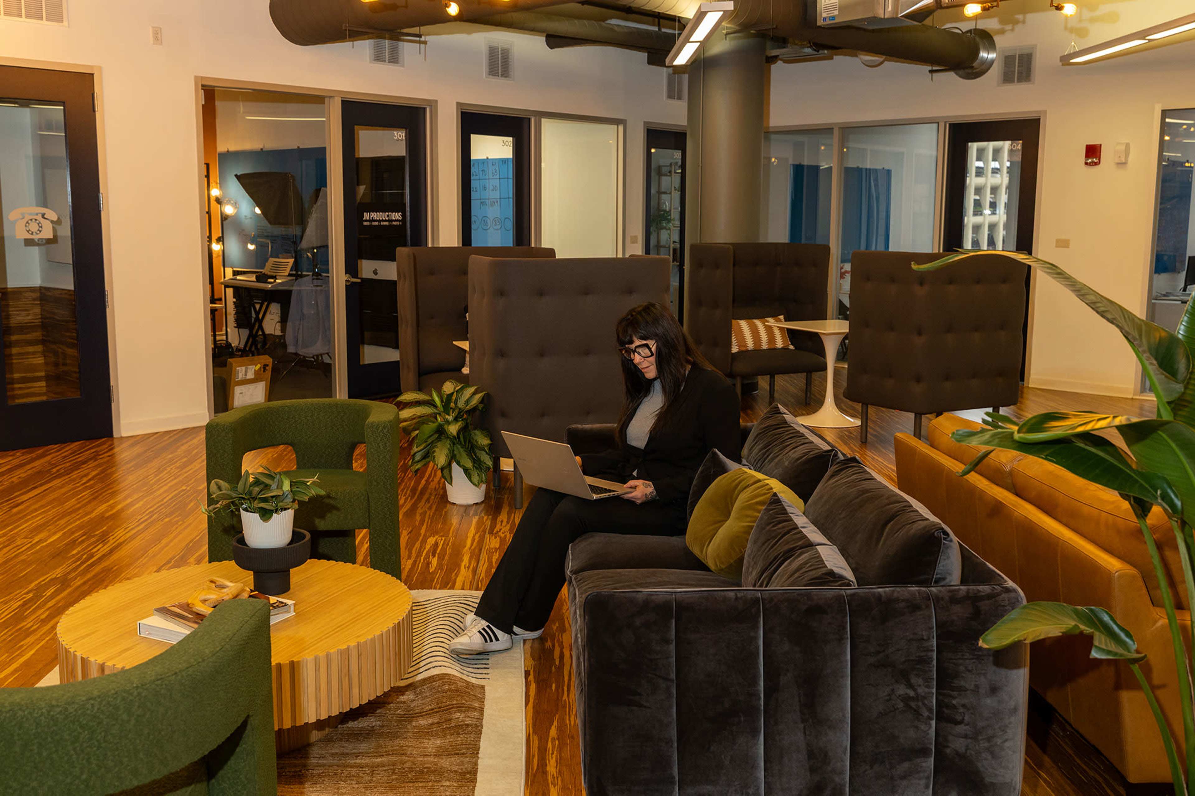 A woman sits at a table using a laptop in a modern office lounge filled with comfortable seating and greenery.