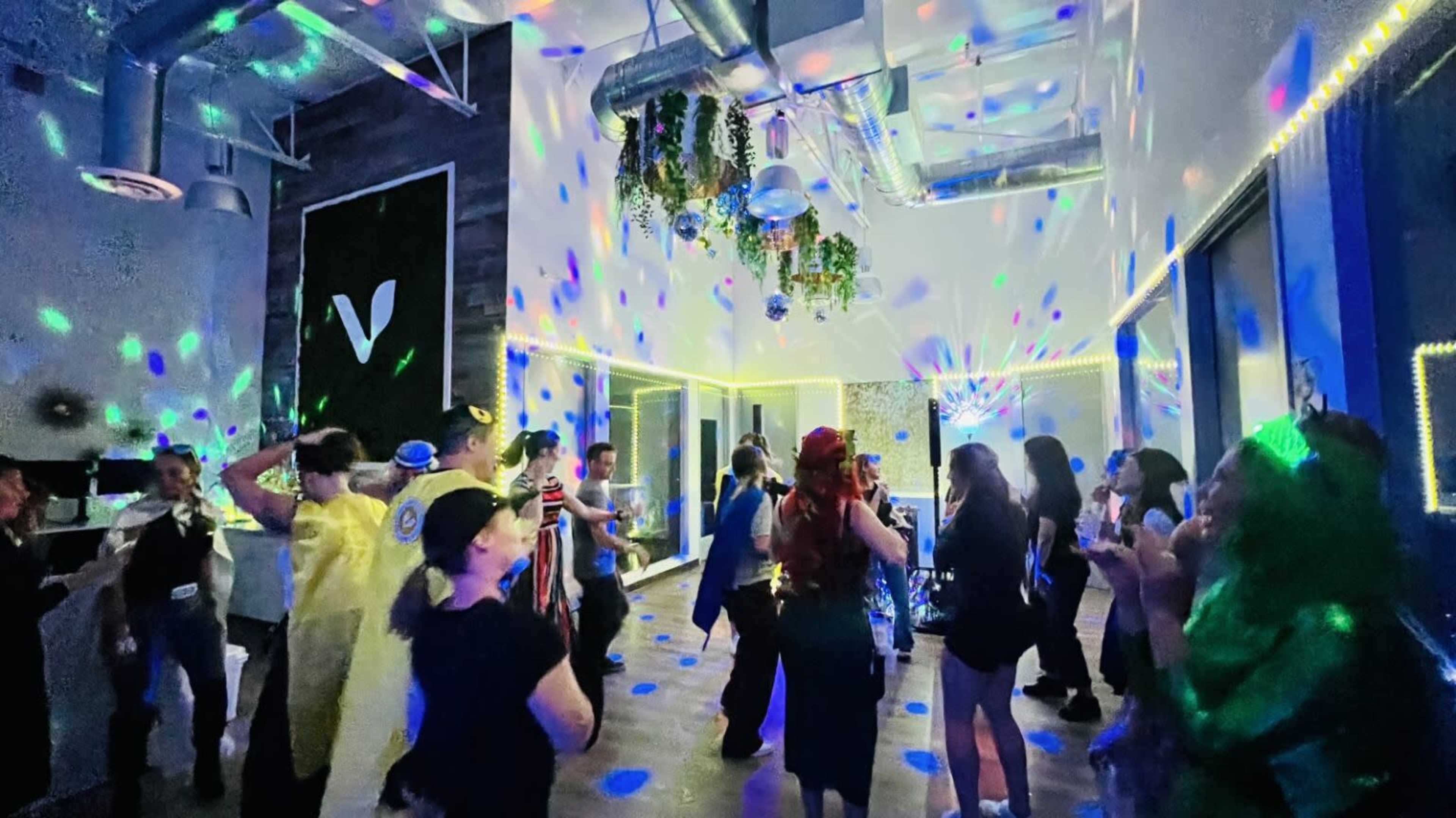 A lively group of people dances under multicolored lights in a spacious indoor venue.