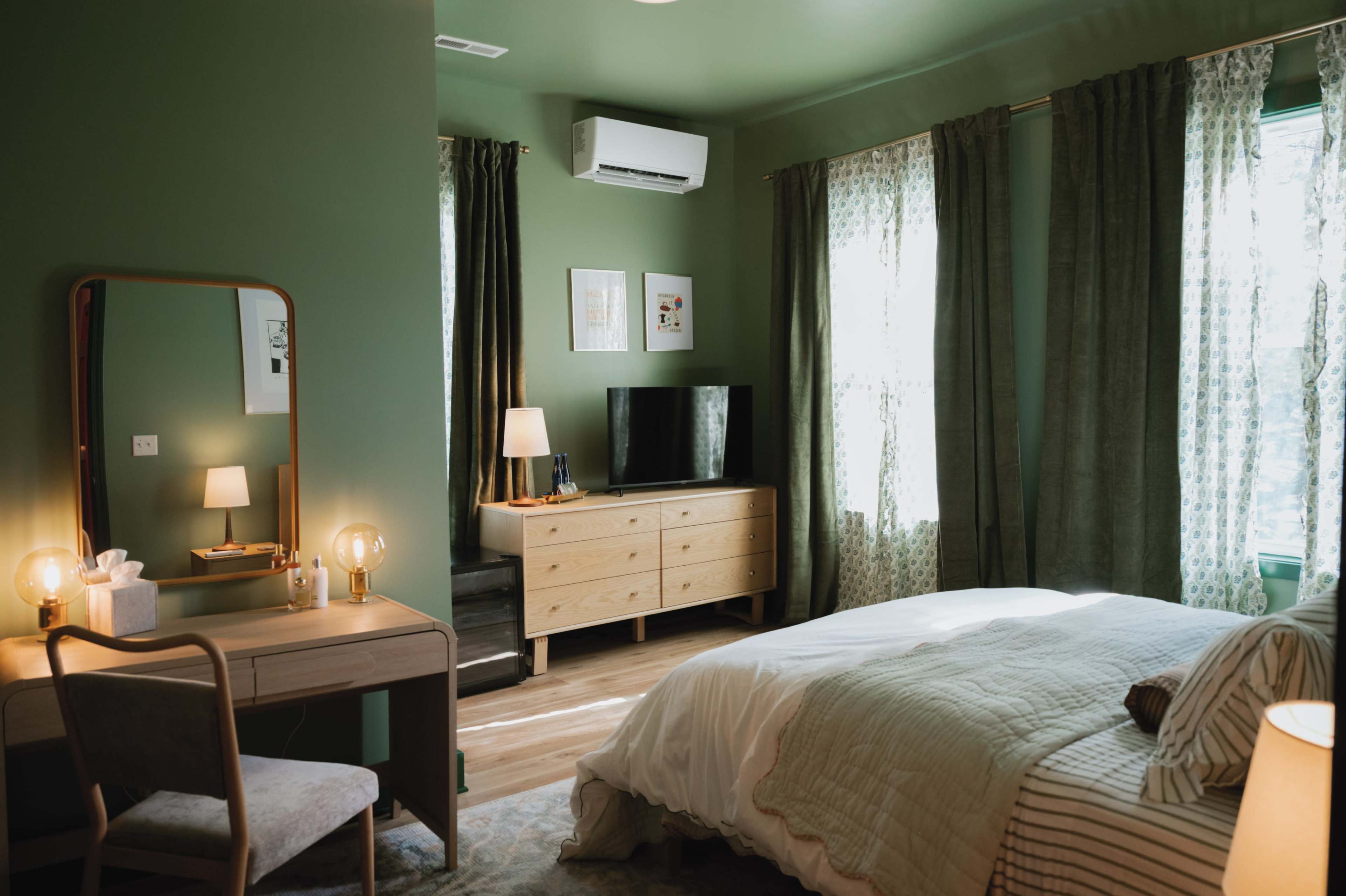 The image shows a stylish bedroom featuring a bed, a wooden dresser with a television, a desk with a chair, and large windows dressed with curtains.