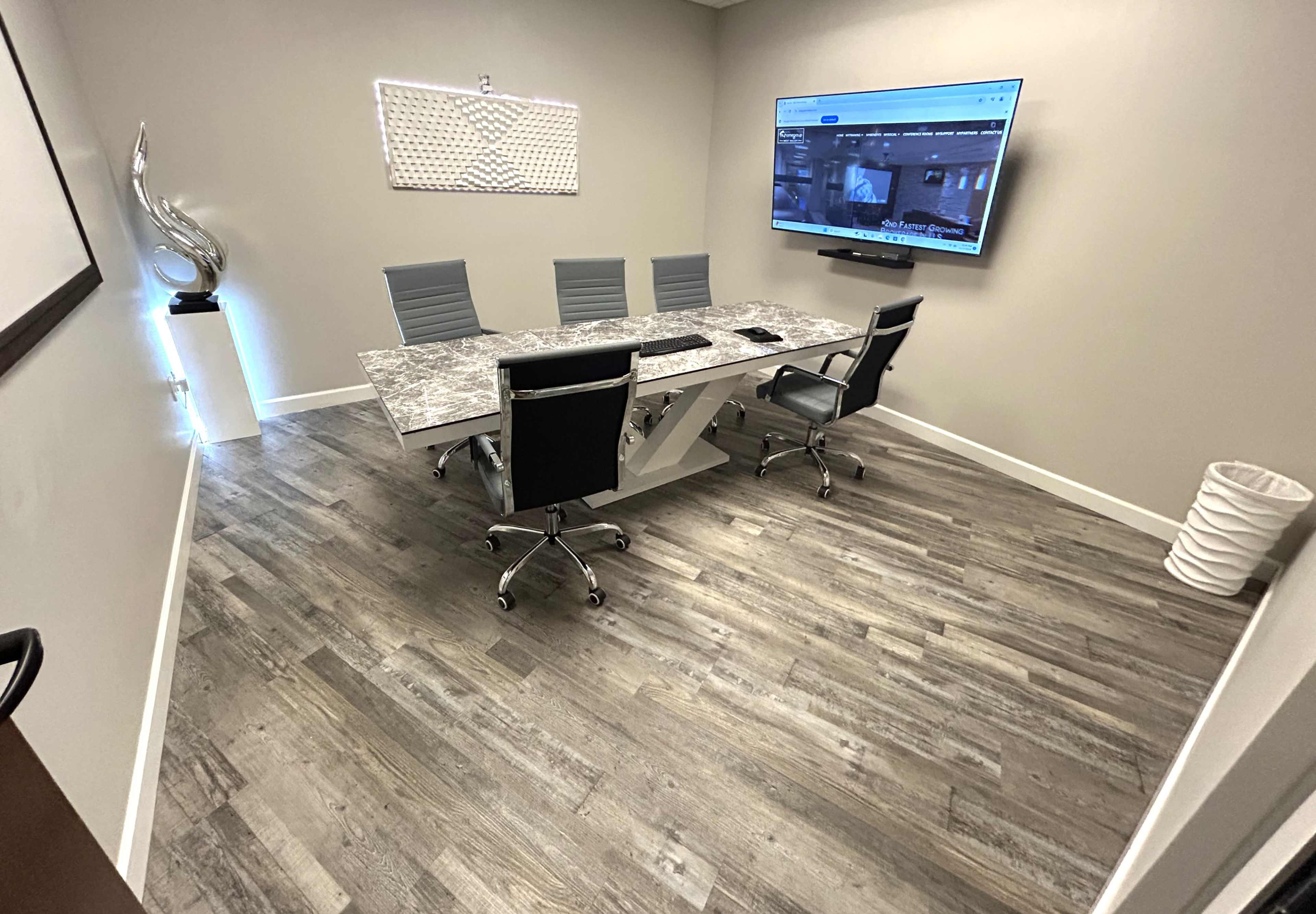 A modern conference room with a large table, four chairs, and a wall-mounted screen displaying a video feed.