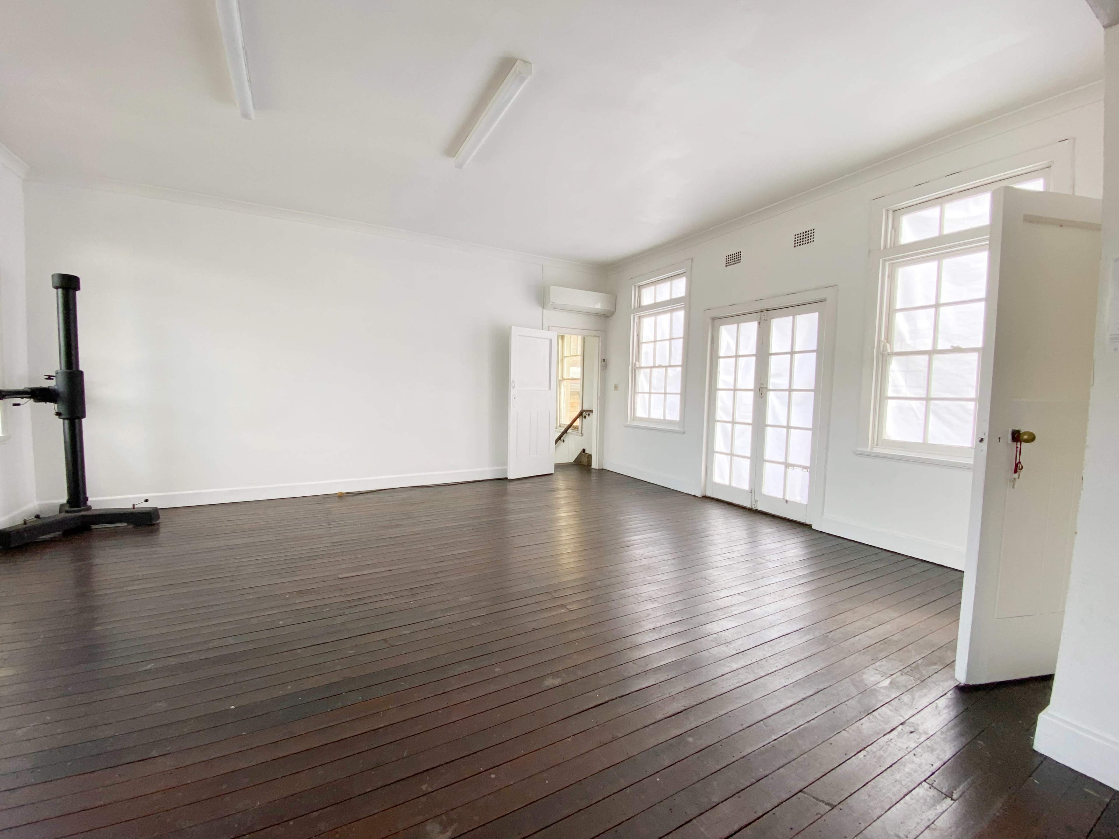 The room features wooden flooring, white walls, and large windows, with a door leading to another area.