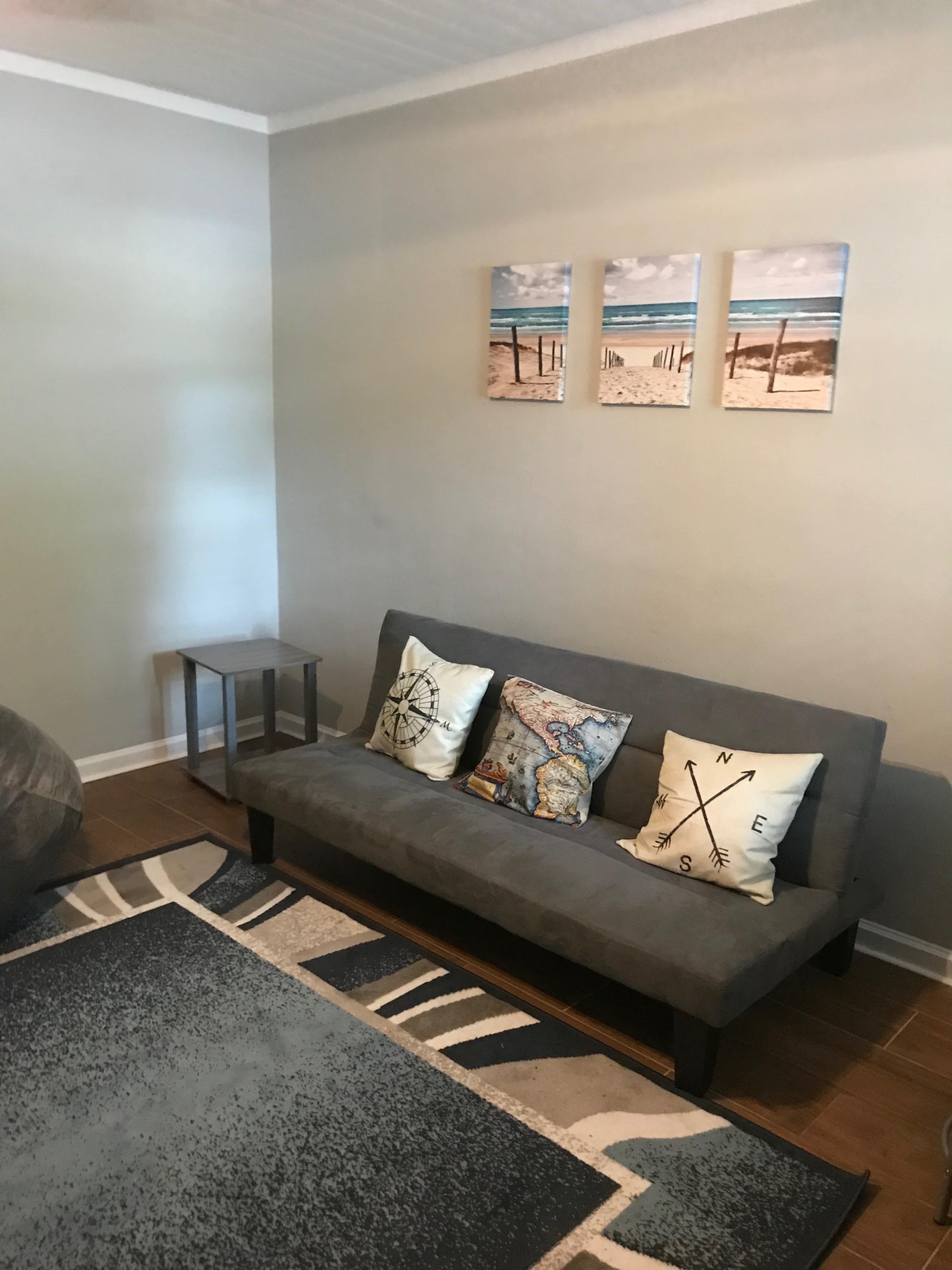 The room features a gray sofa with decorative pillows and a small side table beside it, adorned with beach-themed wall art above.